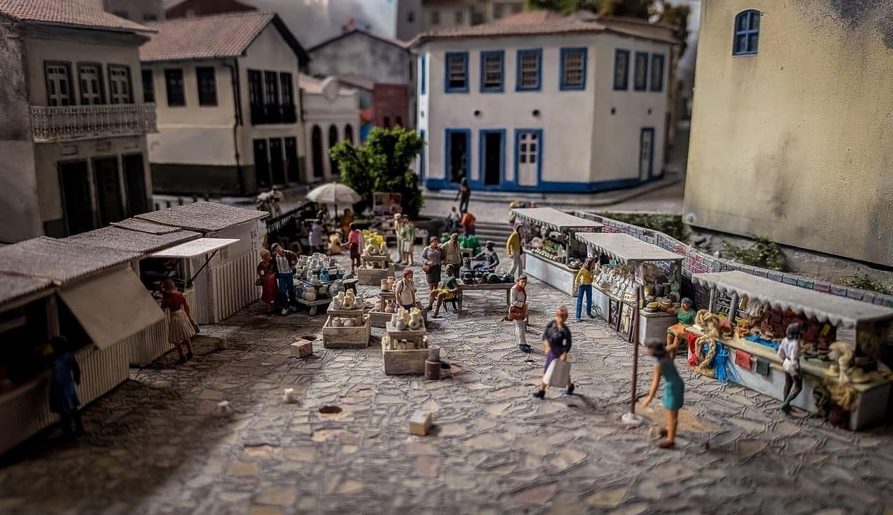 Miniature outdoor market scene on a cobblestone square, with small figurines browsing stalls and tables of goods; a long vendor counter lines the right side, and white buildings with blue-trimmed windows stand in the background.
