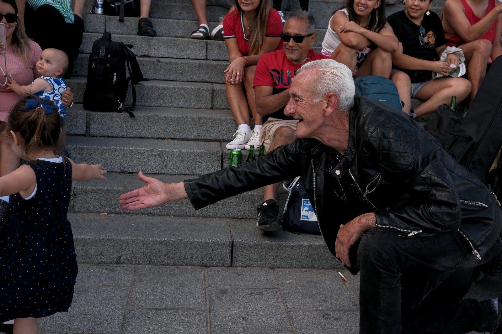 Photo format paysage montrant un vieux monsieur qui dansait à la musique d’un guitariste hors champs et qui se met à genoux pour inviter une petite fille à côté pour une dance, comme un grand père