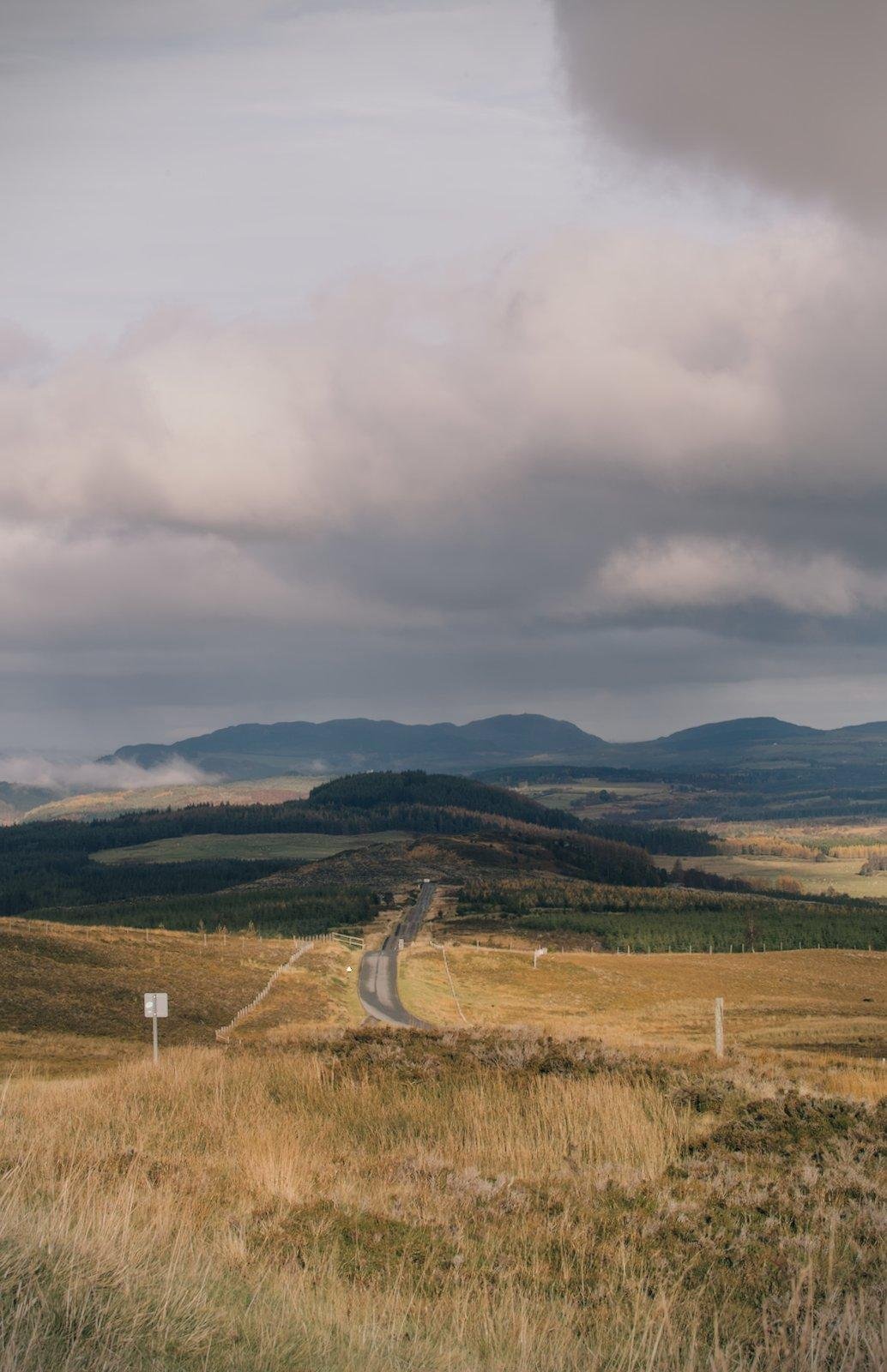 photo format portrait des highlands, avec une route sinueuse au centre, passant au travers de terres dorées pour aller vers des terres verdoyantes au fond, et des monts encore plus loin, sous un ciel couvert d’épais nuages