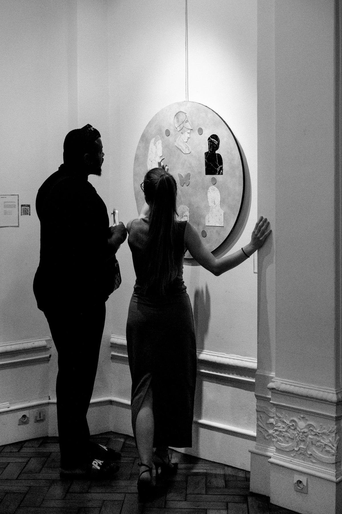 Photo verticale en noir et blanc. Un grand homme bien baraqué regarde une œuvre d’art accrochée au mur, au côté d’une femme toute fine qui explique son travail