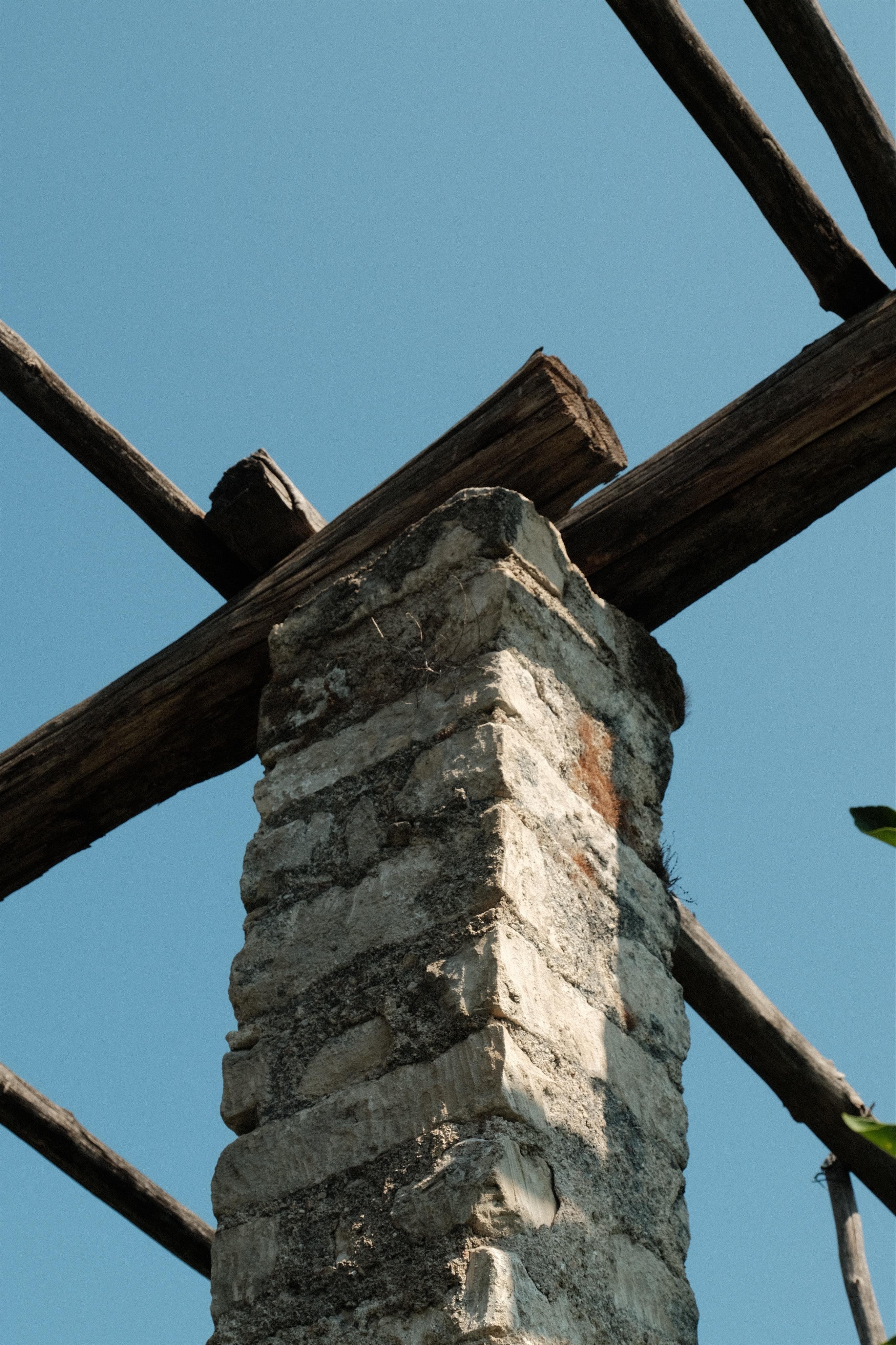 Low-angle view of a weathered stone pillar with rough mortar and intersecting wooden beams overhead, set against a clear blue sky.