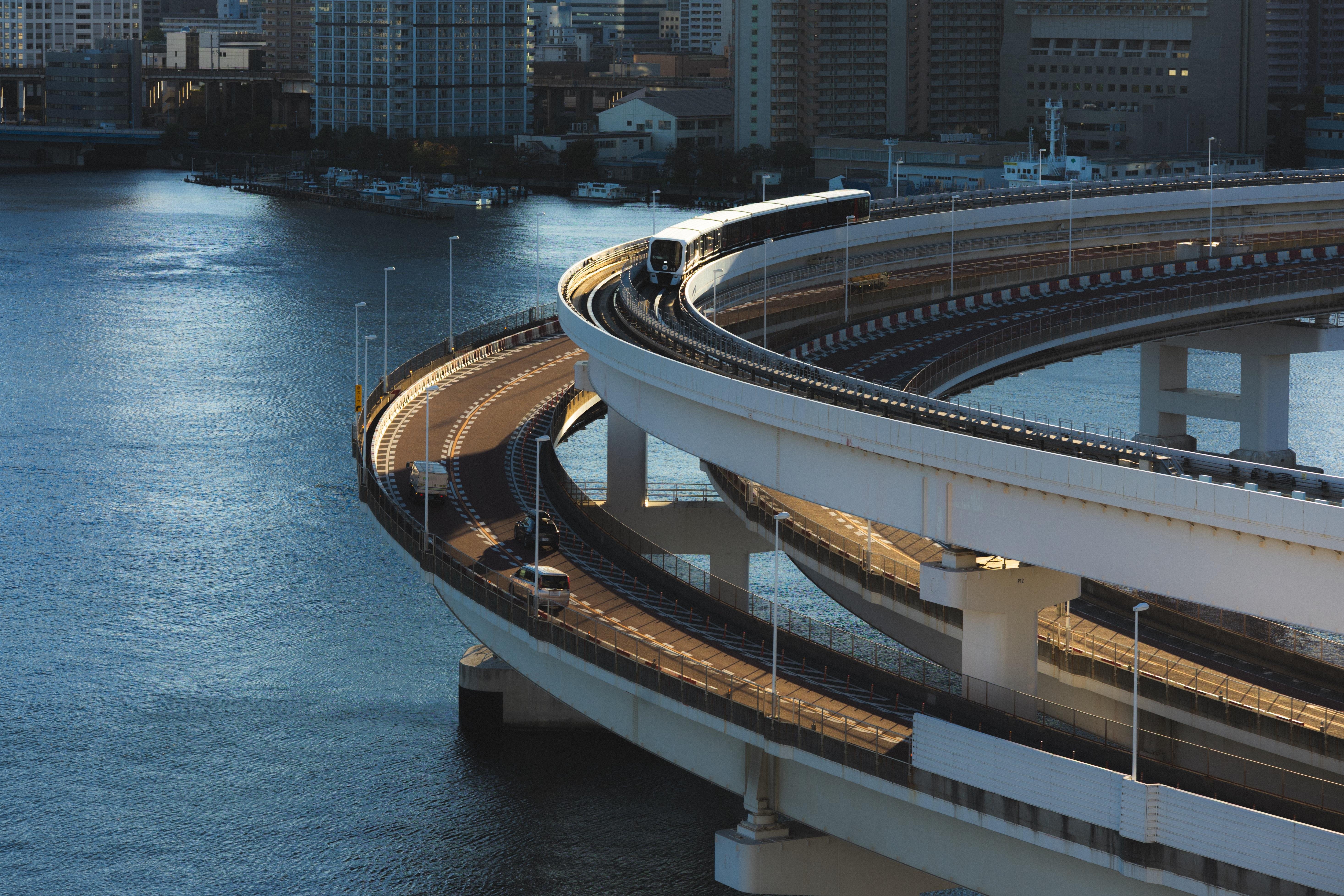Curved highway over water with multiple lanes, elevated train running on upper level, cars on lower levels, surrounded by urban buildings and water reflections.