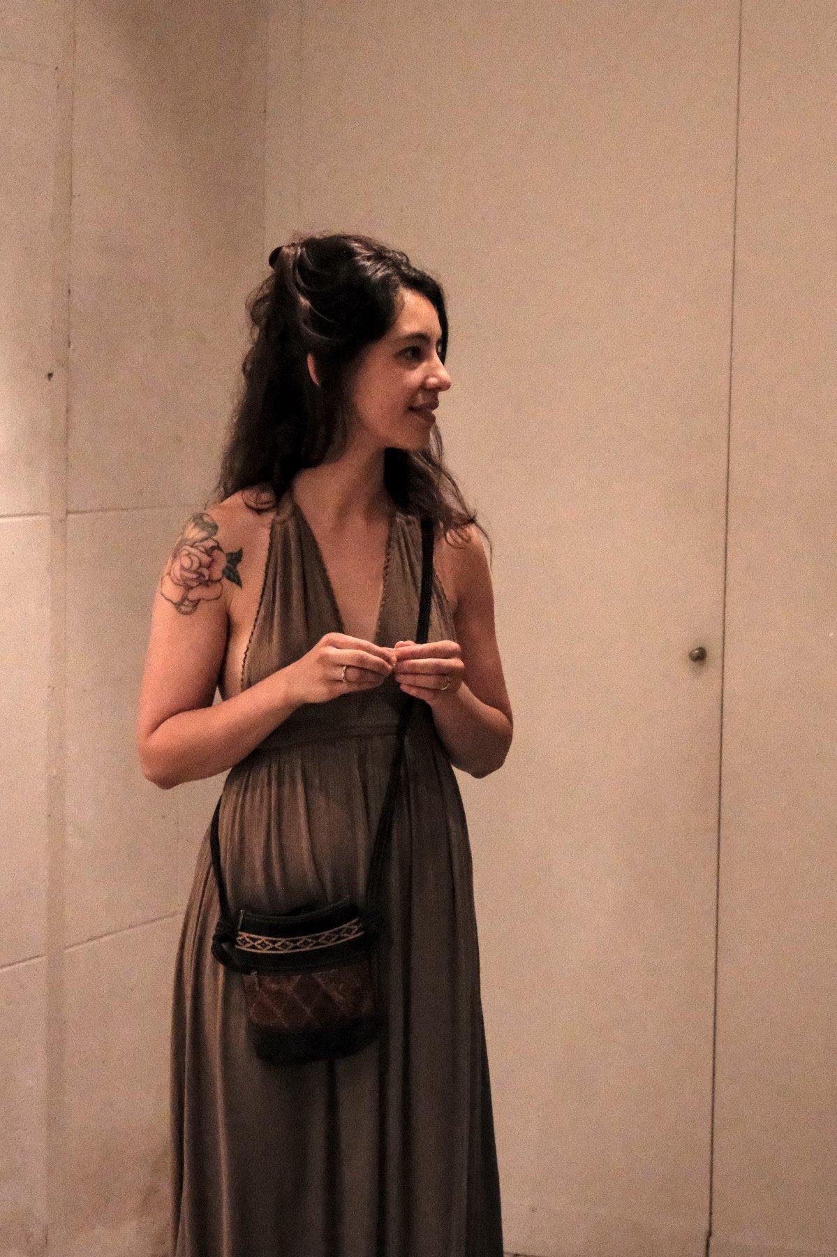 Photo verticale. Une jeune femme au long cheveux brun, en longue robe matton sans manche qui s’attache derrière la nuque, tourne la tête sur sa gauche. Elle semble tenir un truc du bout des doigts des 2 mains