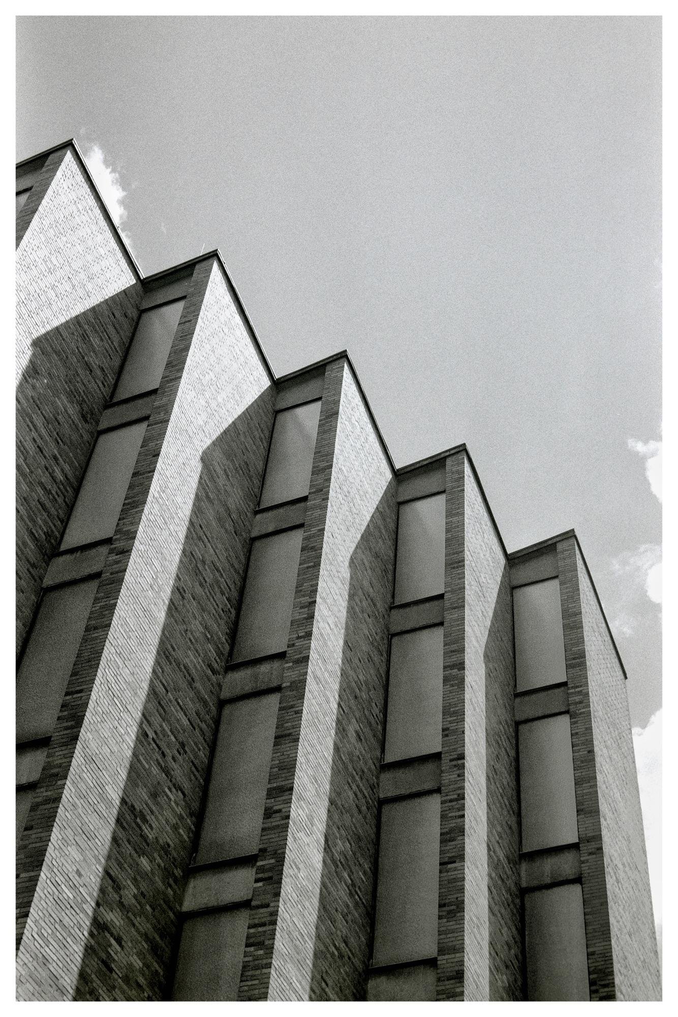 Black and white photo of a tall brick building with a geometric facade, featuring vertical window panels. The building's design creates a pattern of light and shadow across its surface. The sky is visible, with a few clouds at the top corners.