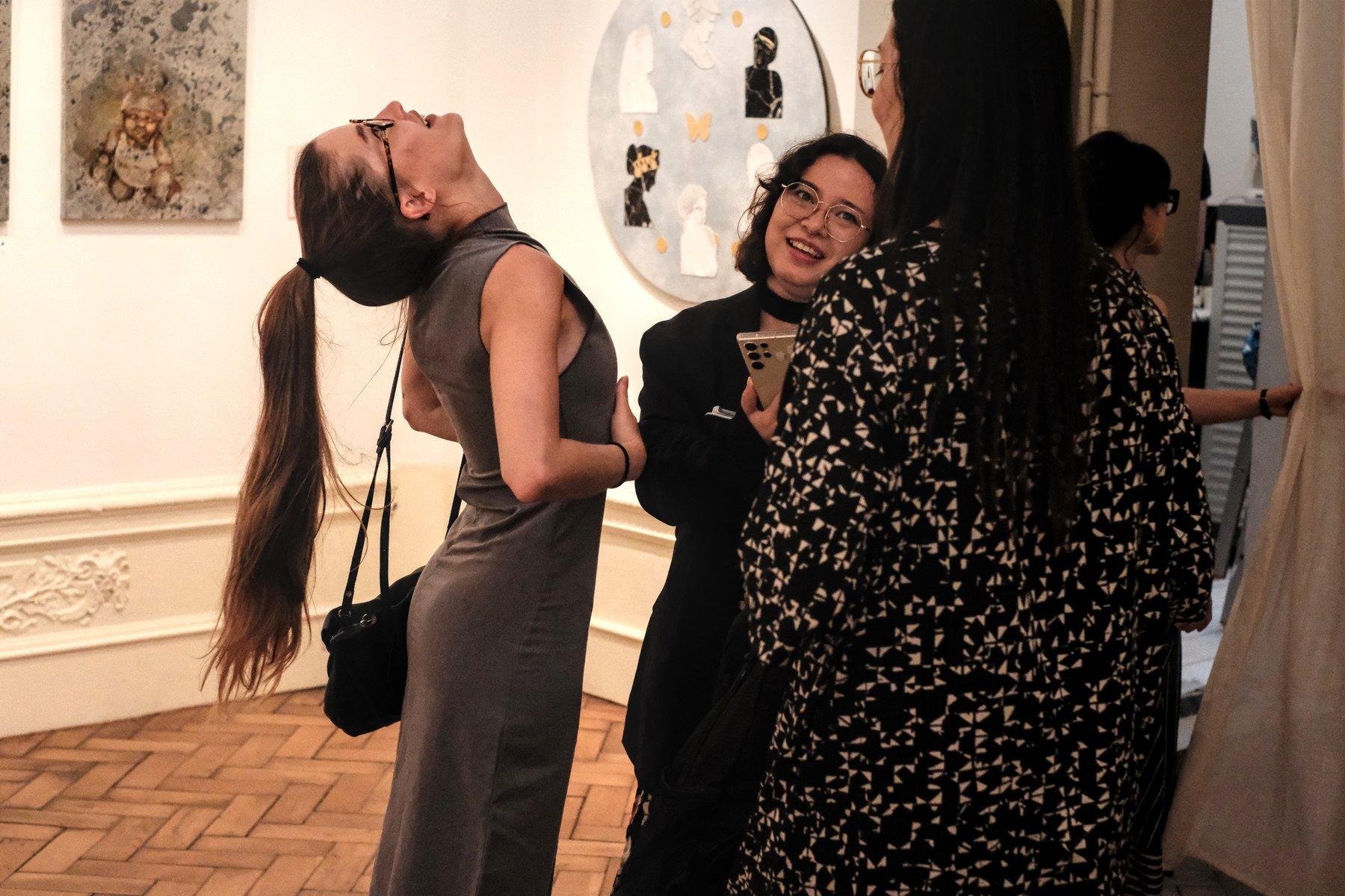 Photo horizontale. 3 jeunes femmes en triangle, discutant au milieu d’une galerie d’art. Celle à gauche, en robe moulante grise, penche la tête en arrière pour regarder le plafond, probablement pour s’étirer le dos. Sa longue queue de cheval reste verticale. Au fond, des œuvres d’art accrochées au mur