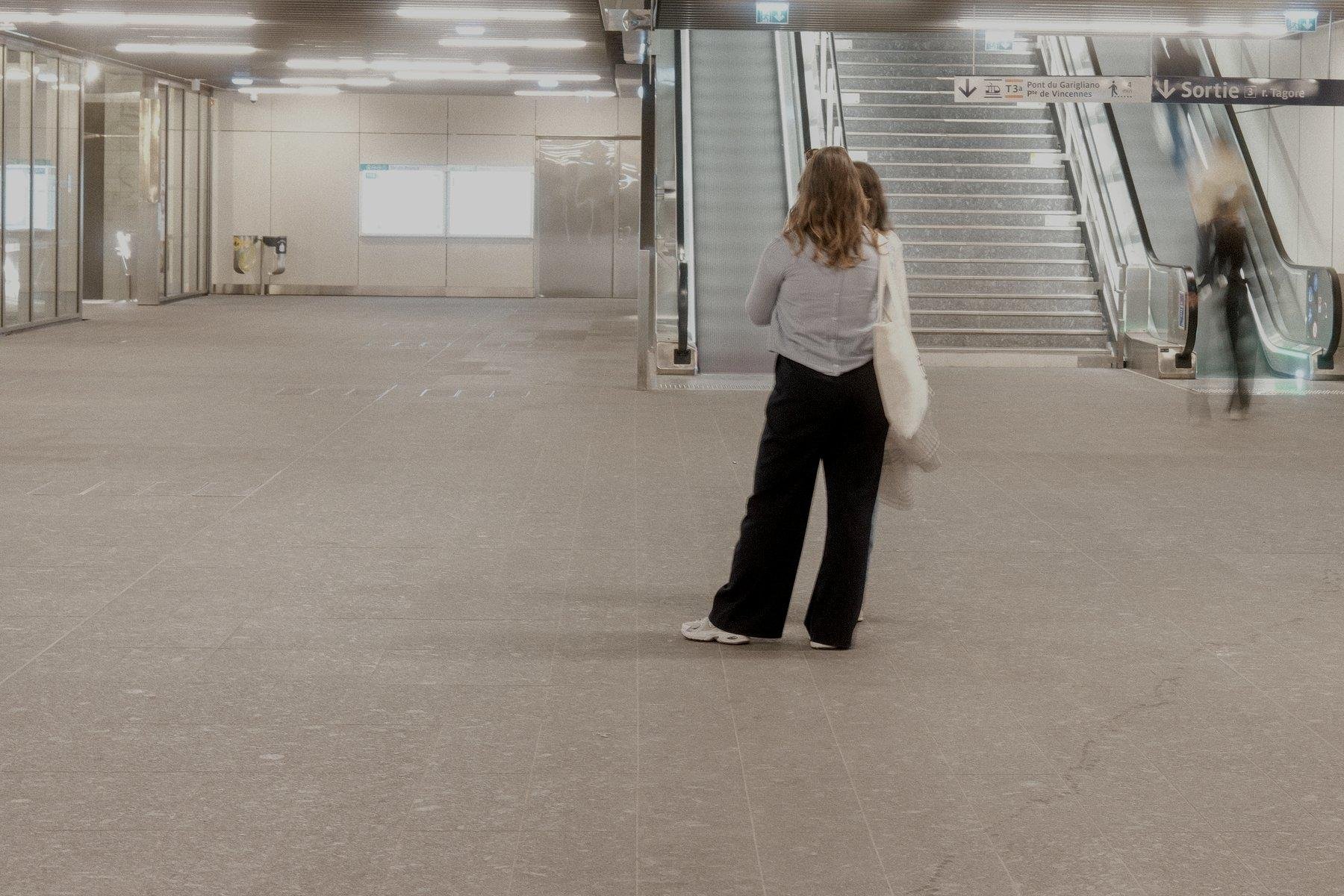 Photo horizontale. Grande salle de passage du métro éclairé au néon. 2 femmes nettes entrain de discuter alors que les passants flou en mouvement sortent de la station par des escalators