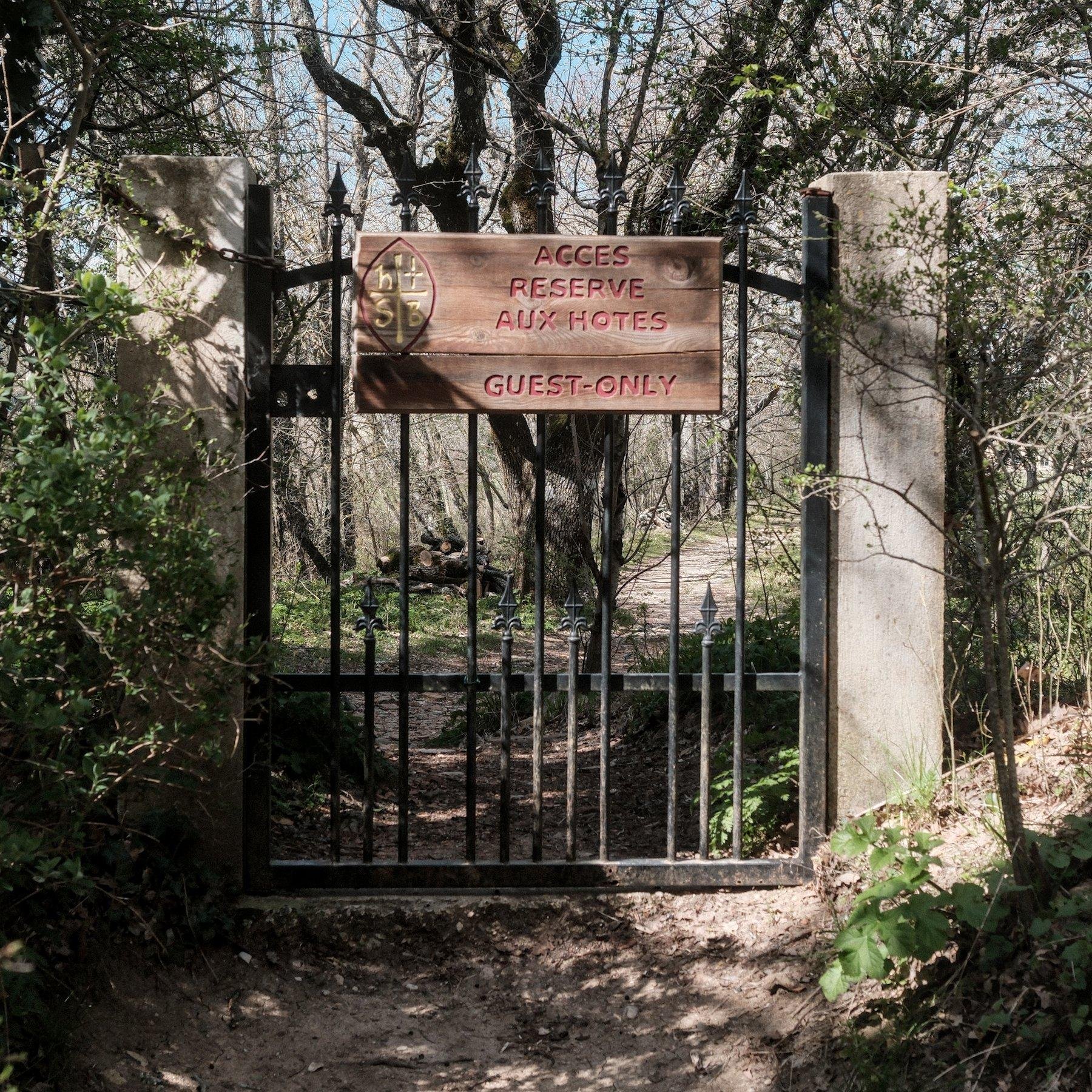 Photo carrée. Un petit portail carré en fer noir sur un chemin de terre, avec une pancarte en bois dessus où il est marqué «&nbsp;Accès réservé aux hôtes - Guest Only&nbsp;»