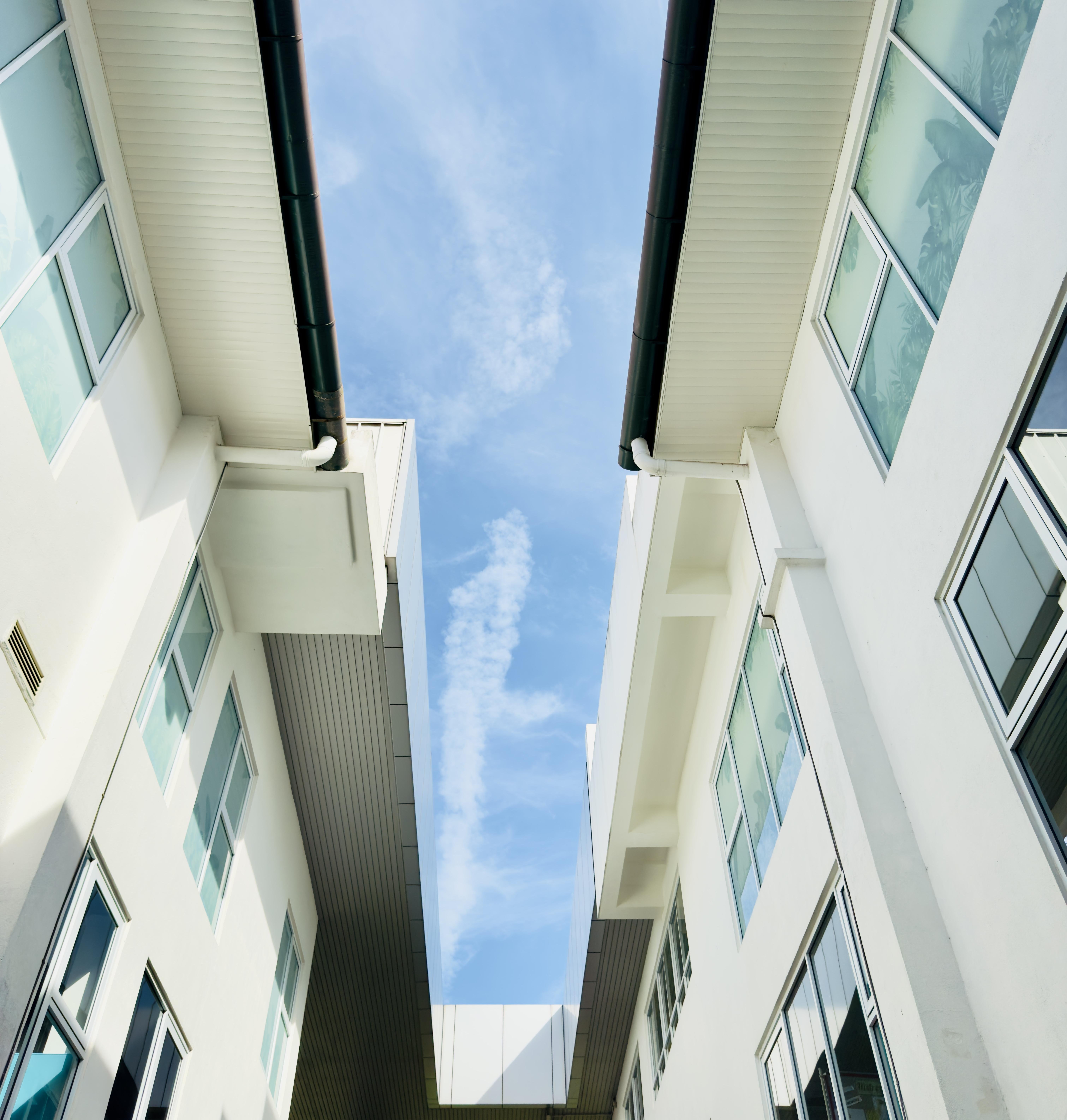 The image shows the facades of two modern buildings with white walls and multiple windows, looking upward toward the sky. The buildings are parallel to one another, creating a narrow space between them. The sky above is blue with some light clouds.