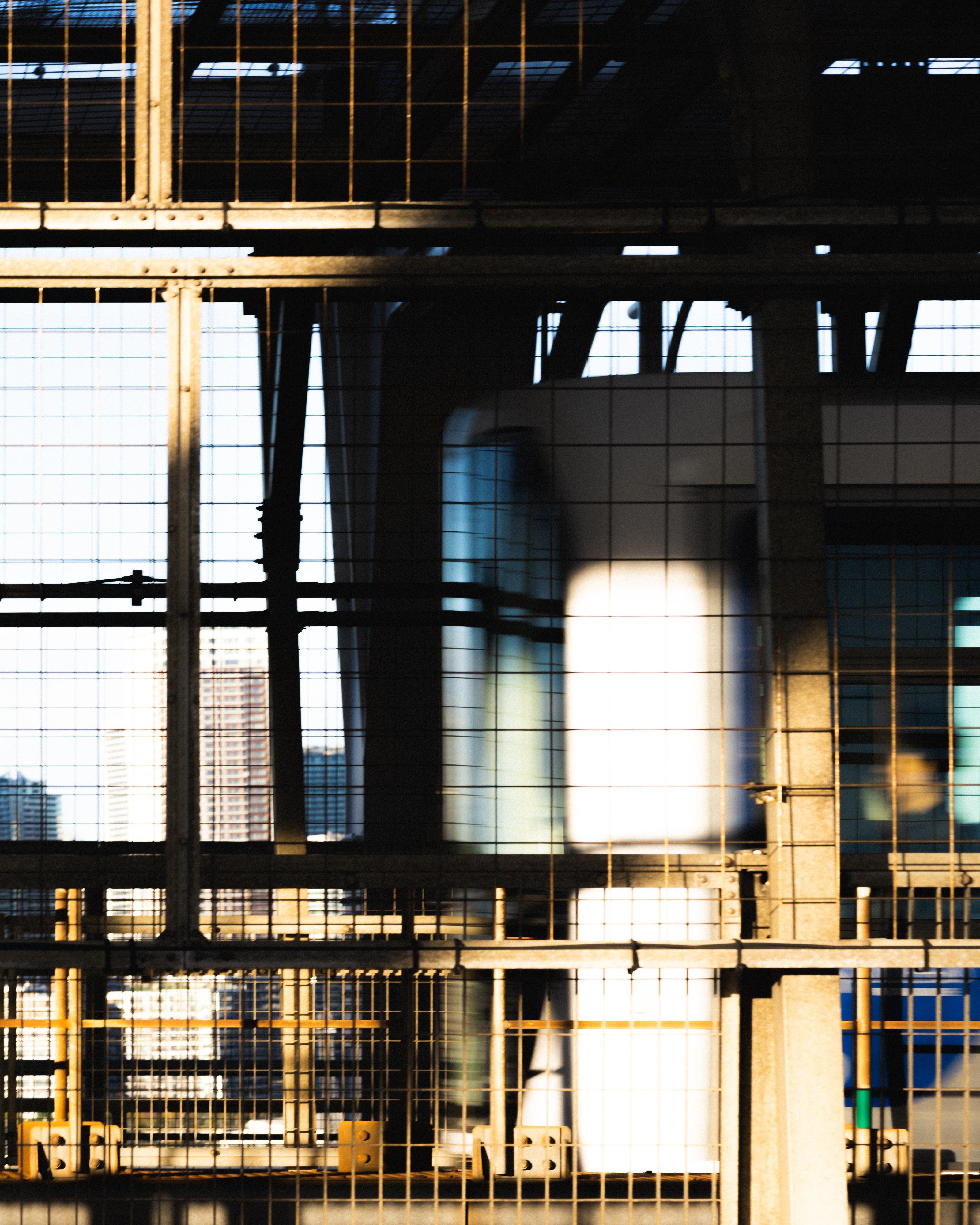 Steel framework with a grid-like pattern dominates the foreground, casting shadows and lines across the image. A blurred background reveals modern building structures with sunlight illuminating parts of the metal and concrete surfaces. The contrast between light and shadow adds depth to the scene.