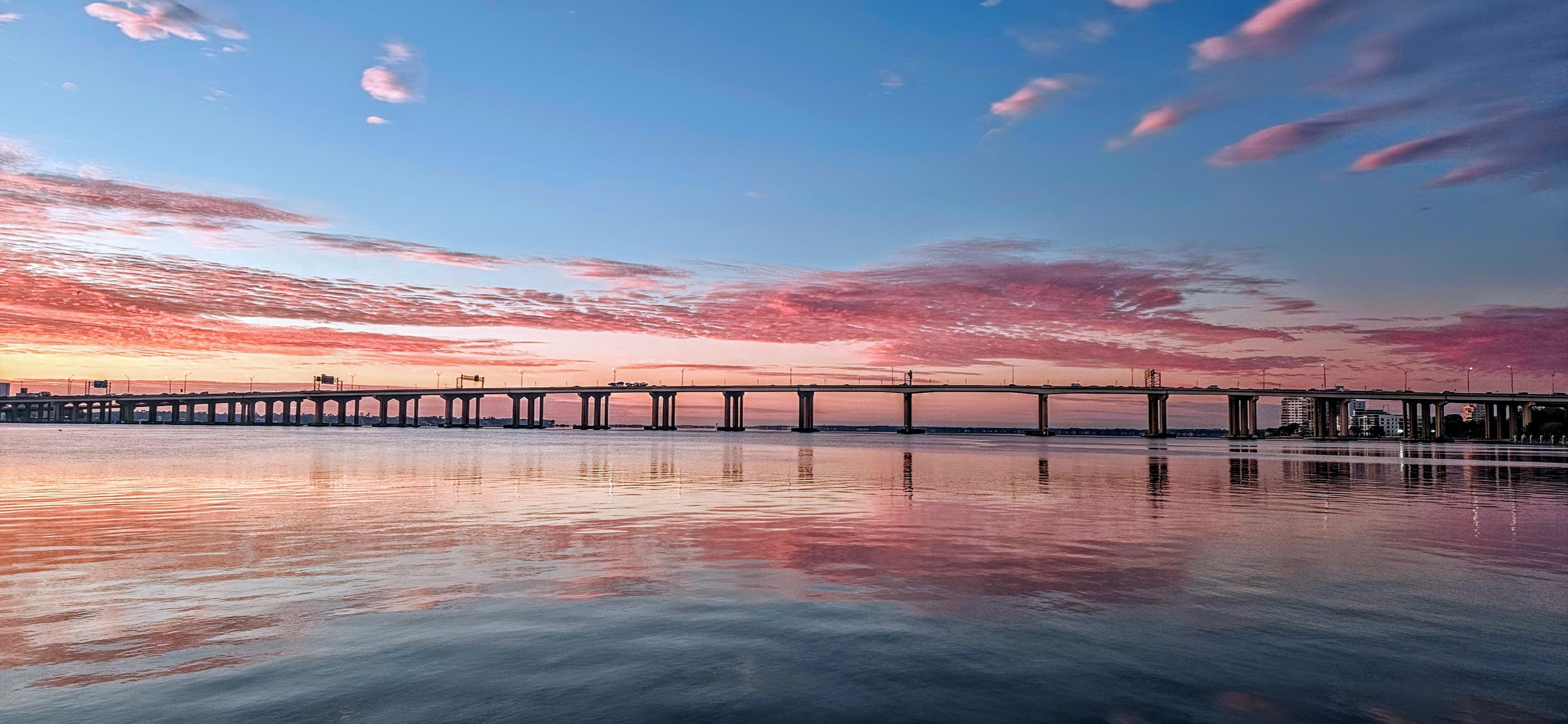 Bridge spans across calm water at sunset, with a sky filled with pink and orange clouds. Reflections of the sky and bridge are visible on the water surface below.
