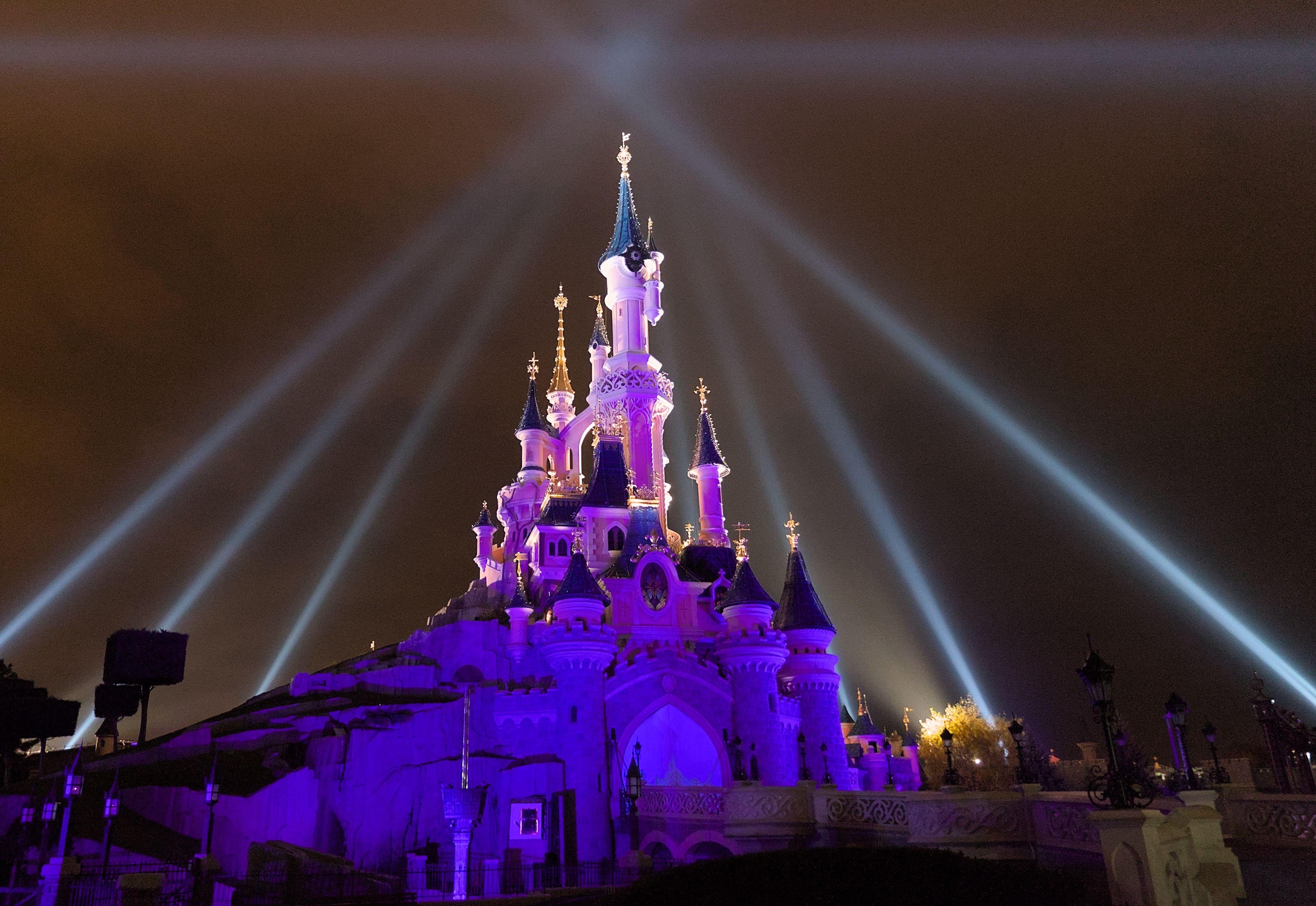 Le château de Disney Paris de nuit, éclairé en teintes violettes et roses, avec les raies de spot de lumière pointant toutes vers un même point au dessus de la plus haute tour