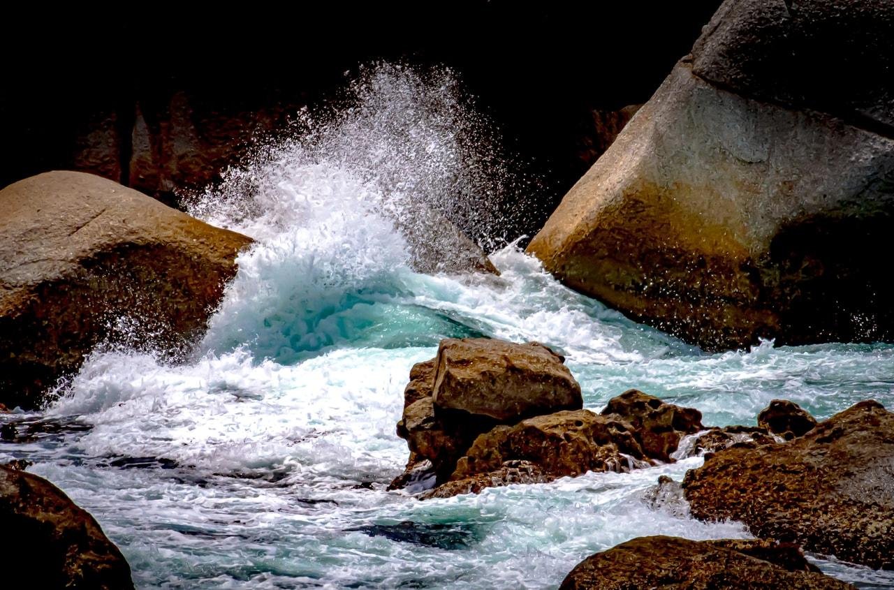 The image shows ocean waves crashing against large rocks. The water appears turbulent, with waves splashing and foaming as they hit the rocky surface. The scene captures the dynamic and powerful interaction between the sea and the rocks.