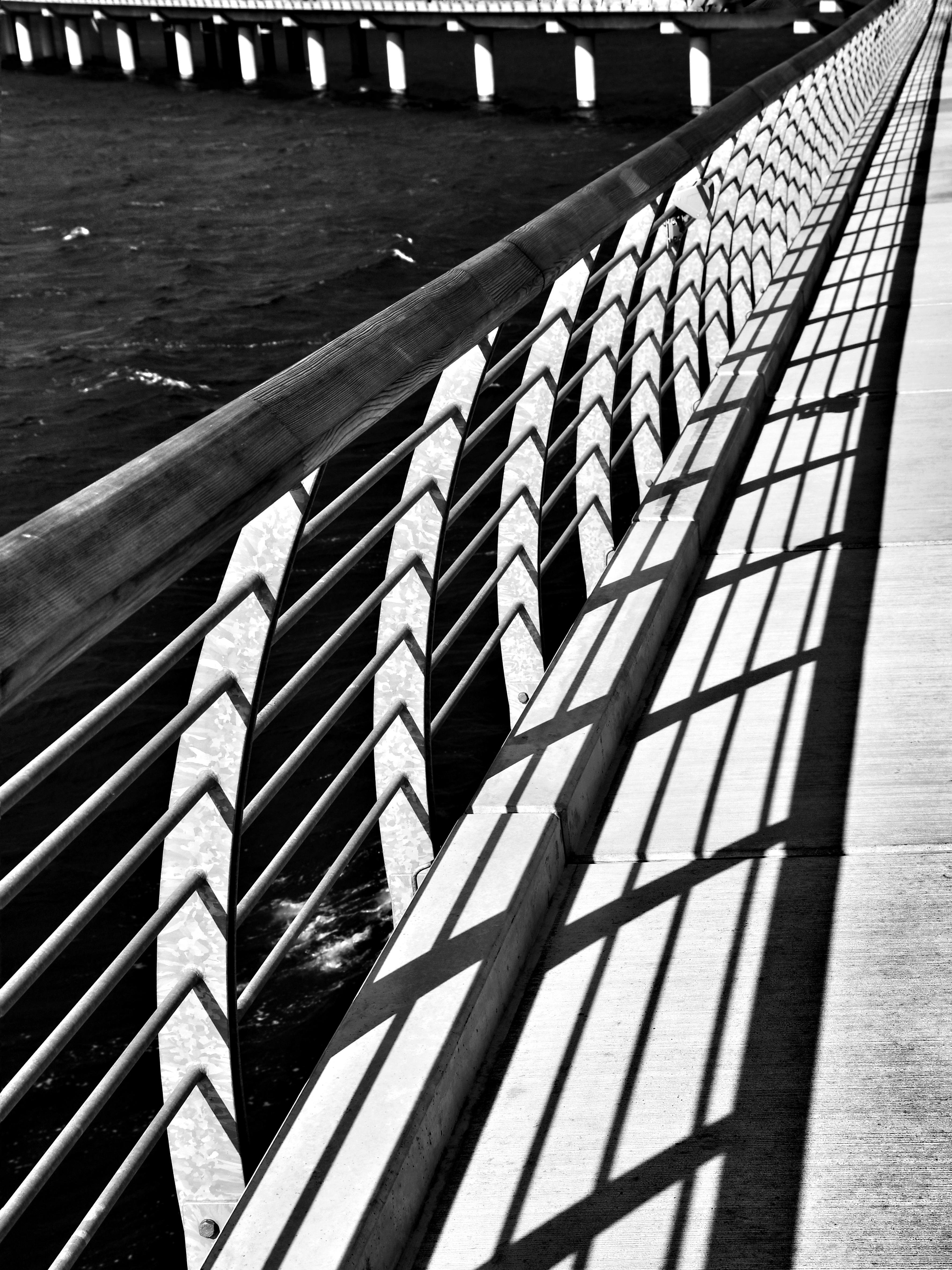 Rails of a bridge create angular shadows on a concrete pathway, forming a distinctive pattern. Below, water is visible with ripples, while a series of white columns support another part of the bridge in the background. The image is in black and white, emphasizing contrast.
###
Brückengeländer werfen eckige Schatten auf einen Betonweg und bilden ein markantes Muster. Darunter sind Wasserwellen zu sehen, während im Hintergrund eine Reihe weißer Säulen einen anderen Teil der Brücke stützen. Das Bild ist schwarz-weiß und betont den Kontrast.