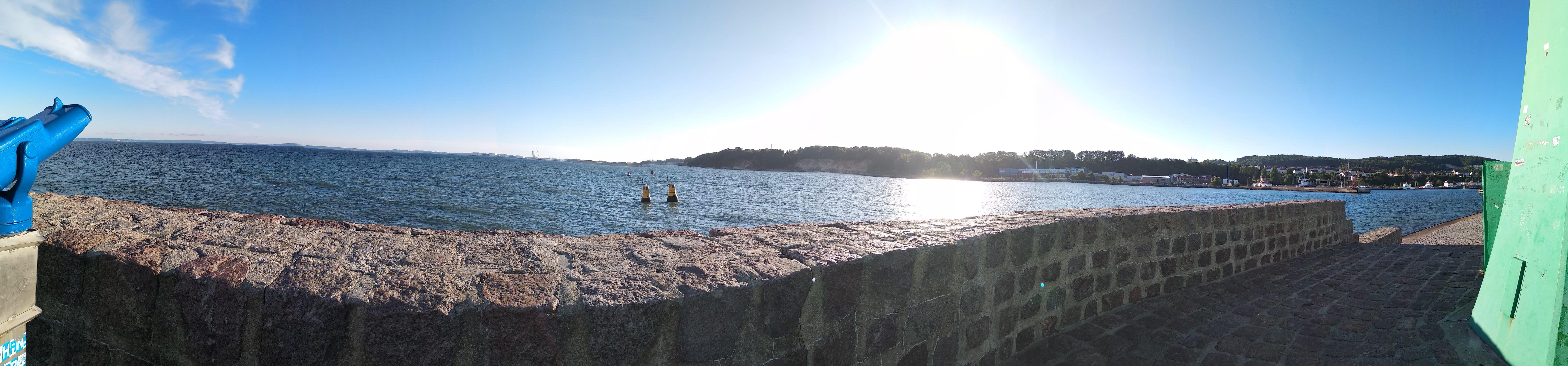 Panoramic view of a coastline with a stone wall in the foreground. Two people are standing in the water near the shore. The sun is low in the sky, creating a bright glare over the water. To the left, a blue coin-operated viewer is visible, while the right side shows part of a green structure. Hills and scattered buildings are seen in the distance on the opposite shore.