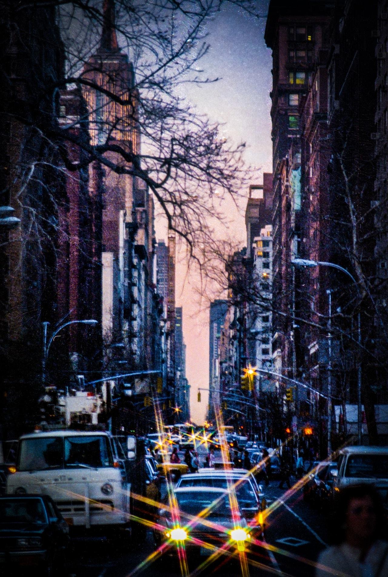 A city street at dusk, framed by tall buildings on both sides. A large building with a pointed top is visible in the distance. Leafless tree branches are silhouetted against the sky. Traffic fills the street, with headlights creating starburst effects. The sky has a gradient from pink to blue as evening approaches.