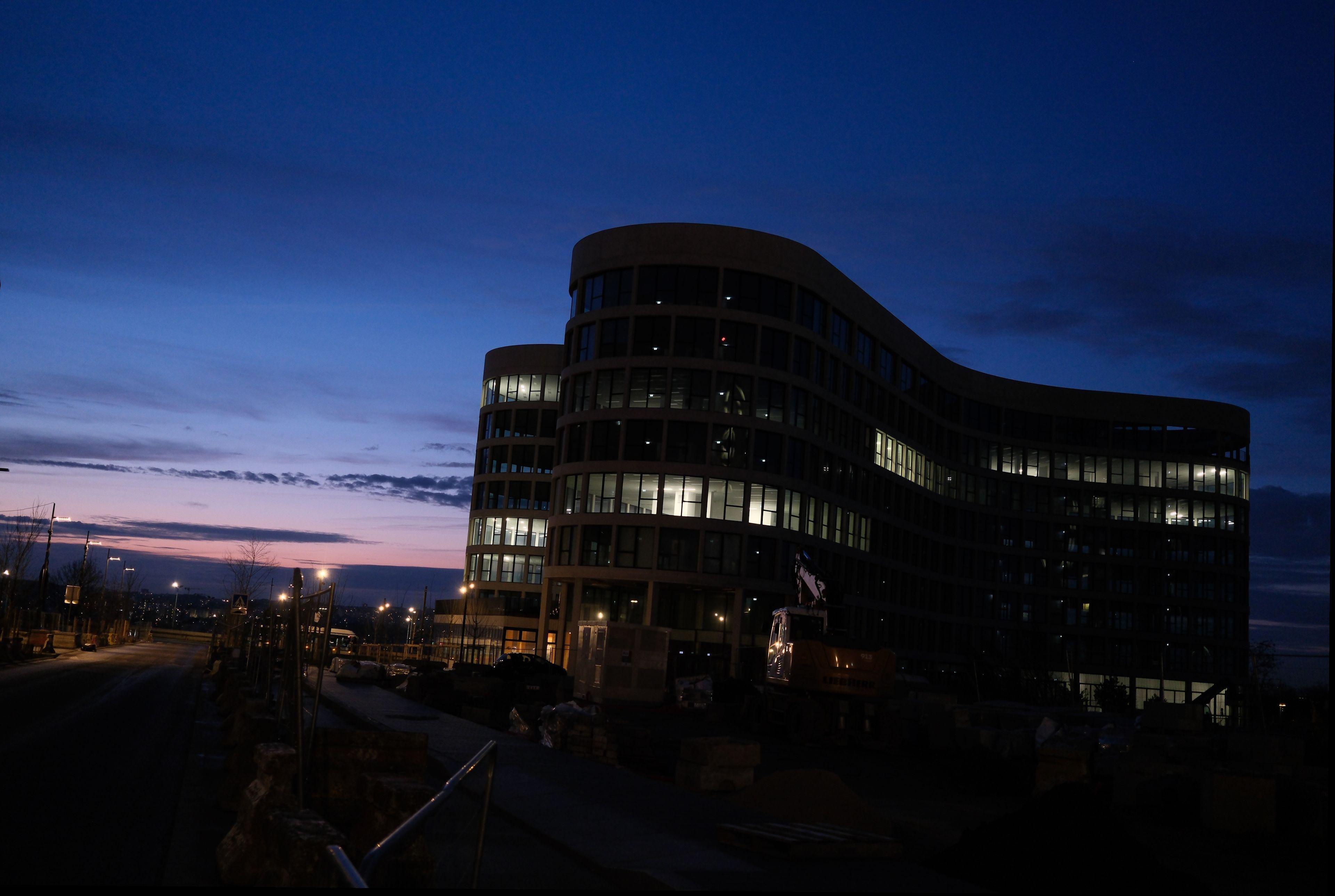 Photo paysage d’un bâtiment à la forme particulière, tout en courbes, très moderne, sur fond de ciel aux couleurs du crépuscule, bleu nuit en haut à droite vers le rose orange en bas à gauche.