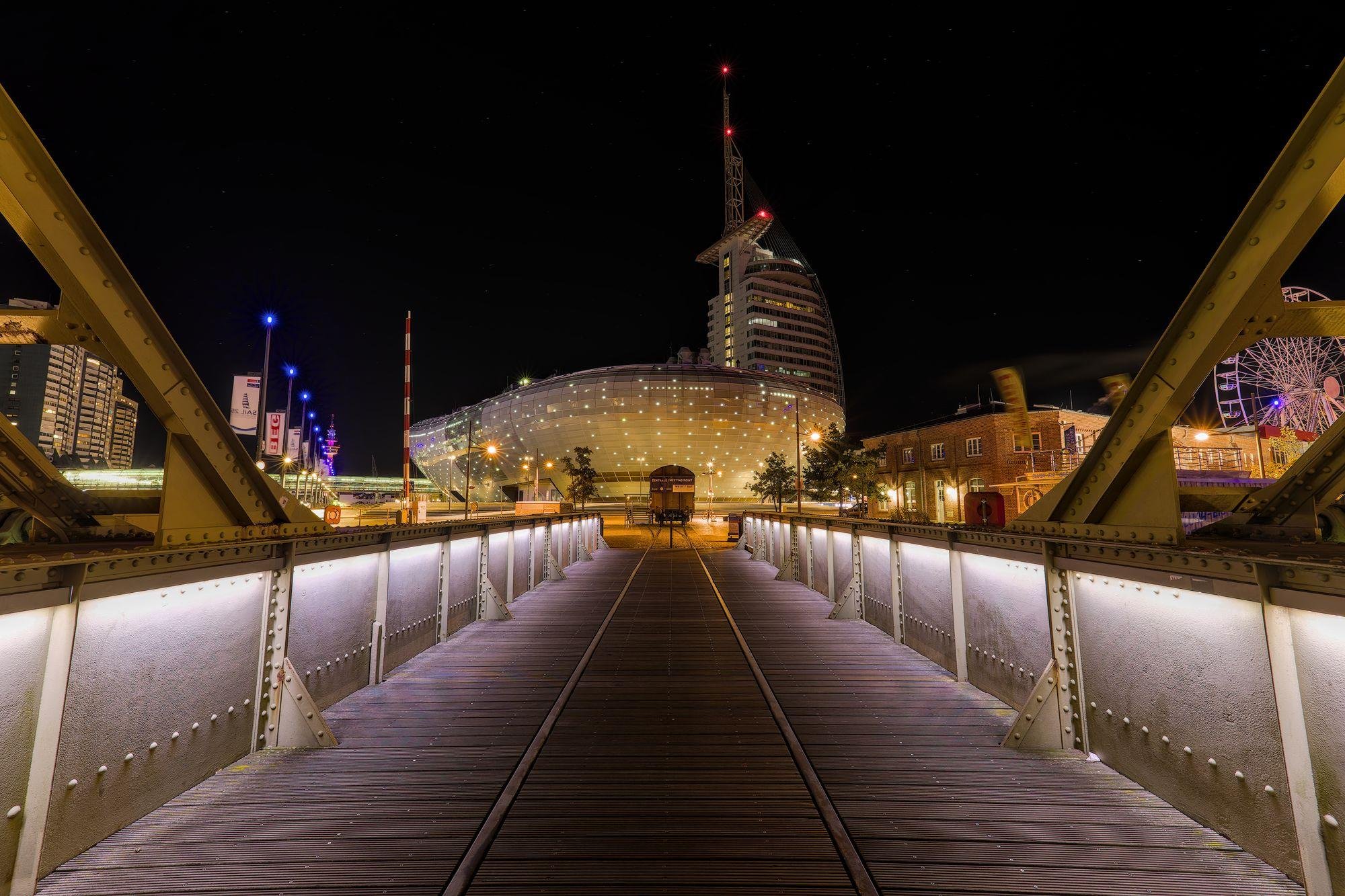 Night view from the center of a metal truss bridge with wooden deck and embedded rail tracks leading to a lit modern curved building with a tower and red warning lights; streetlights and buildings line both sides, with a ferris wheel visible on the right.