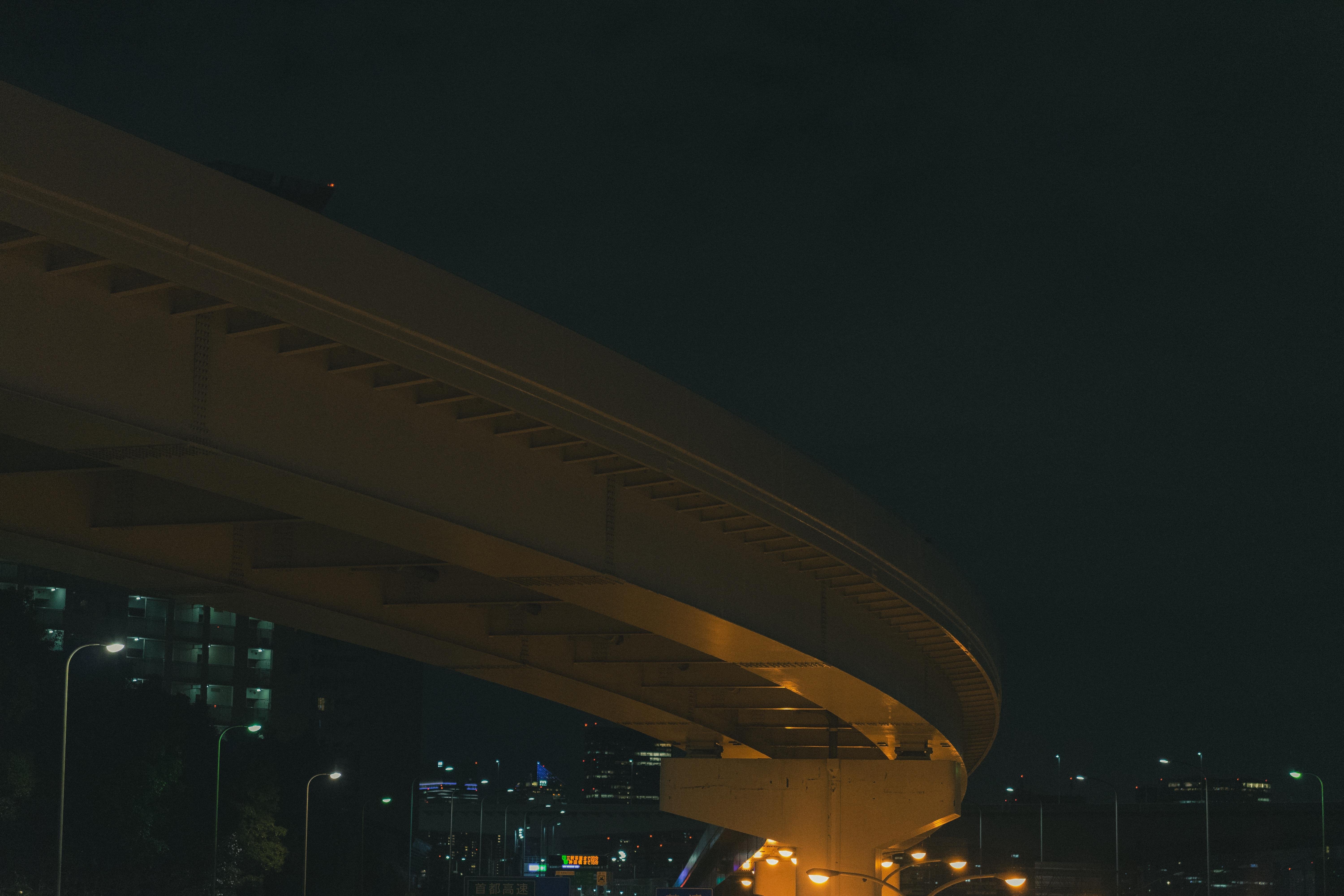 The image shows an elevated highway or bridge at night. It's lit with streetlights, and you can see part of a cityscape in the distance. The scene is dark with a focus on the curve of the road.