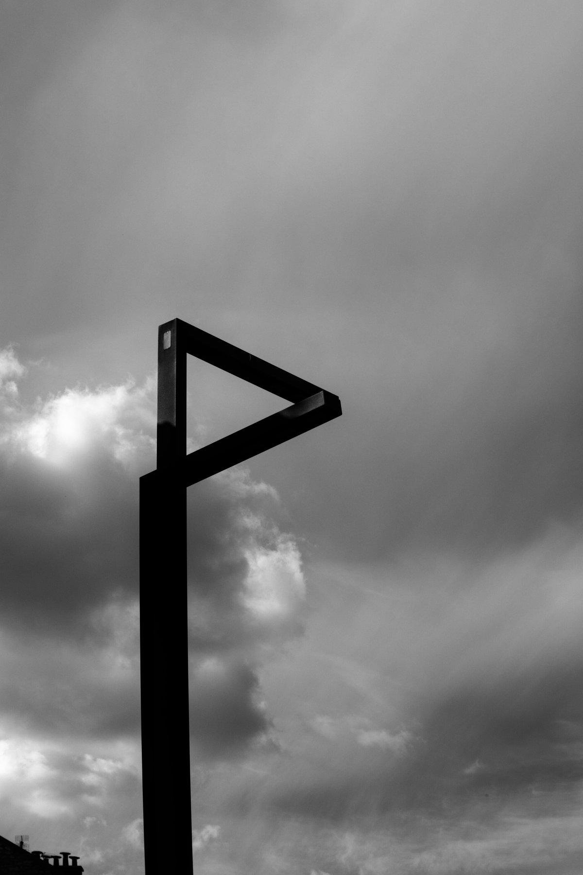 Photo verticale en noir et blanc. 2 lampadaires au design très moderne, en angle droit, perpendiculaires l’un par rapport à l’autre, mais la perspective forme un triangle de Penrose avec