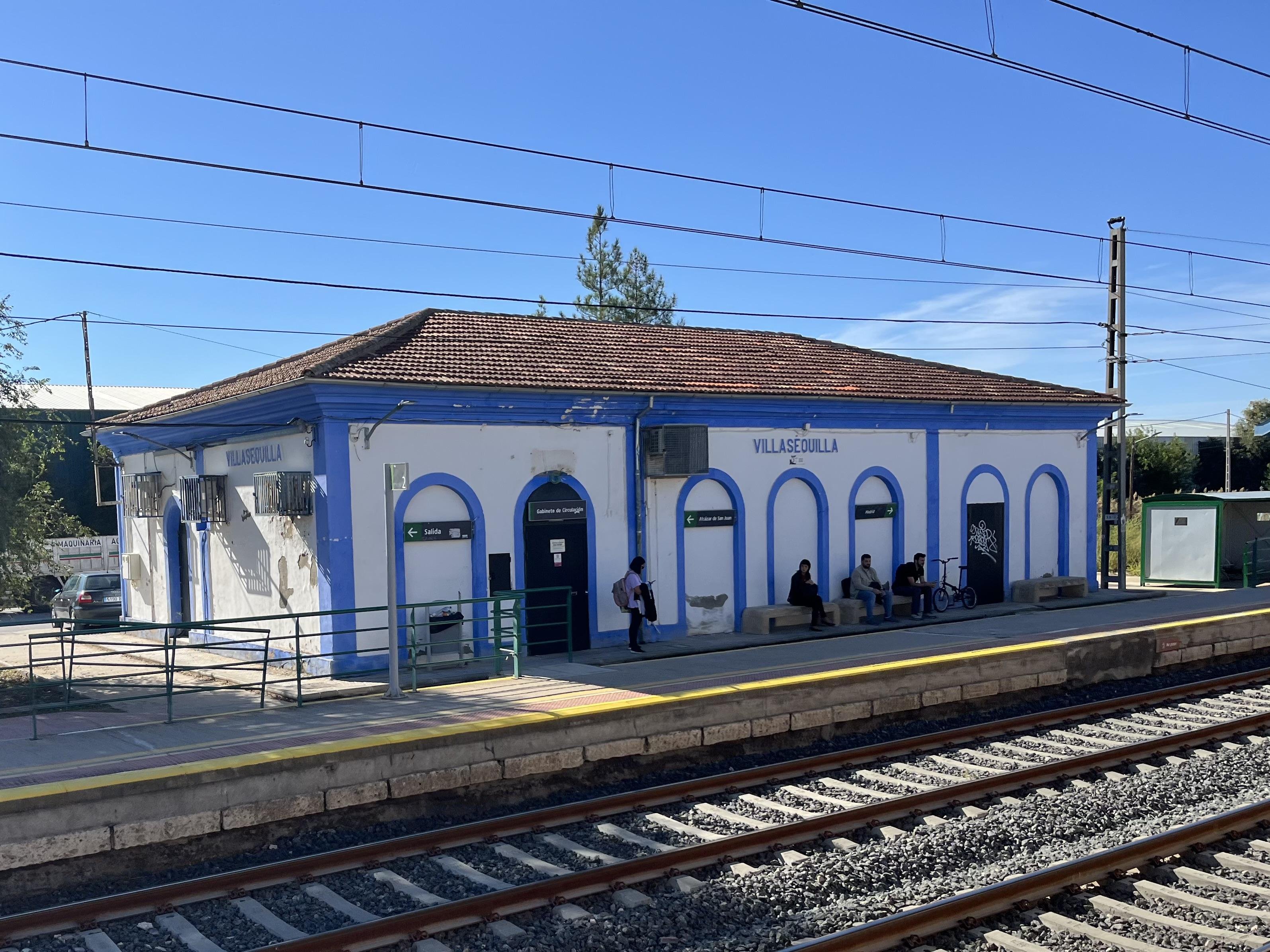 The image shows a small train station with a building labeled "Villasequilla." There are railroad tracks in the foreground and several people sitting on benches outside the building. The architecture is simple with a white facade and blue trim. Weather is clear with a blue sky.
