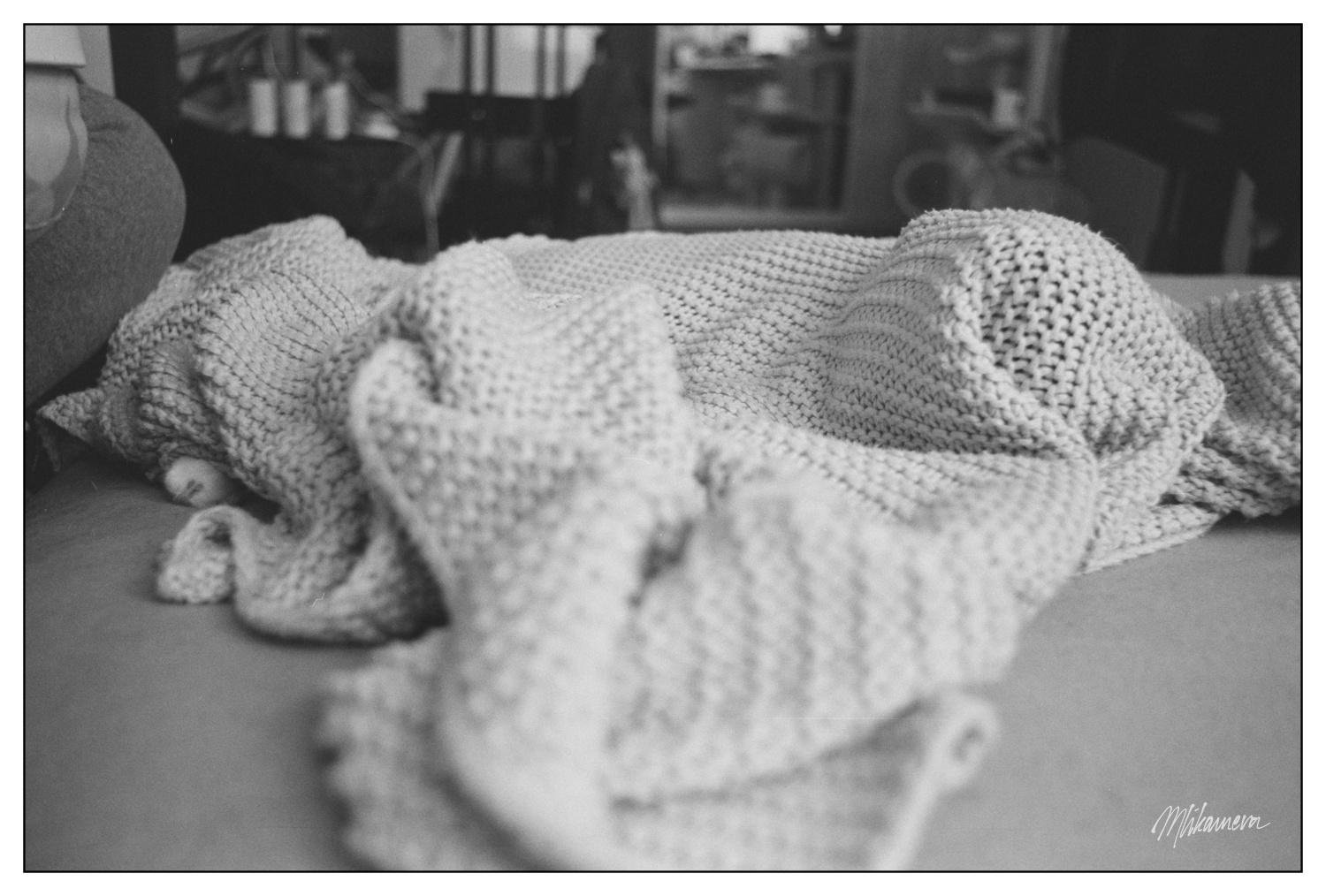 A monochrome photo of a knitted blanket on a bed with roughly cylindrical shape underneath. Upon closer inspection, there is a white paw 
visible on the left side of the shape.