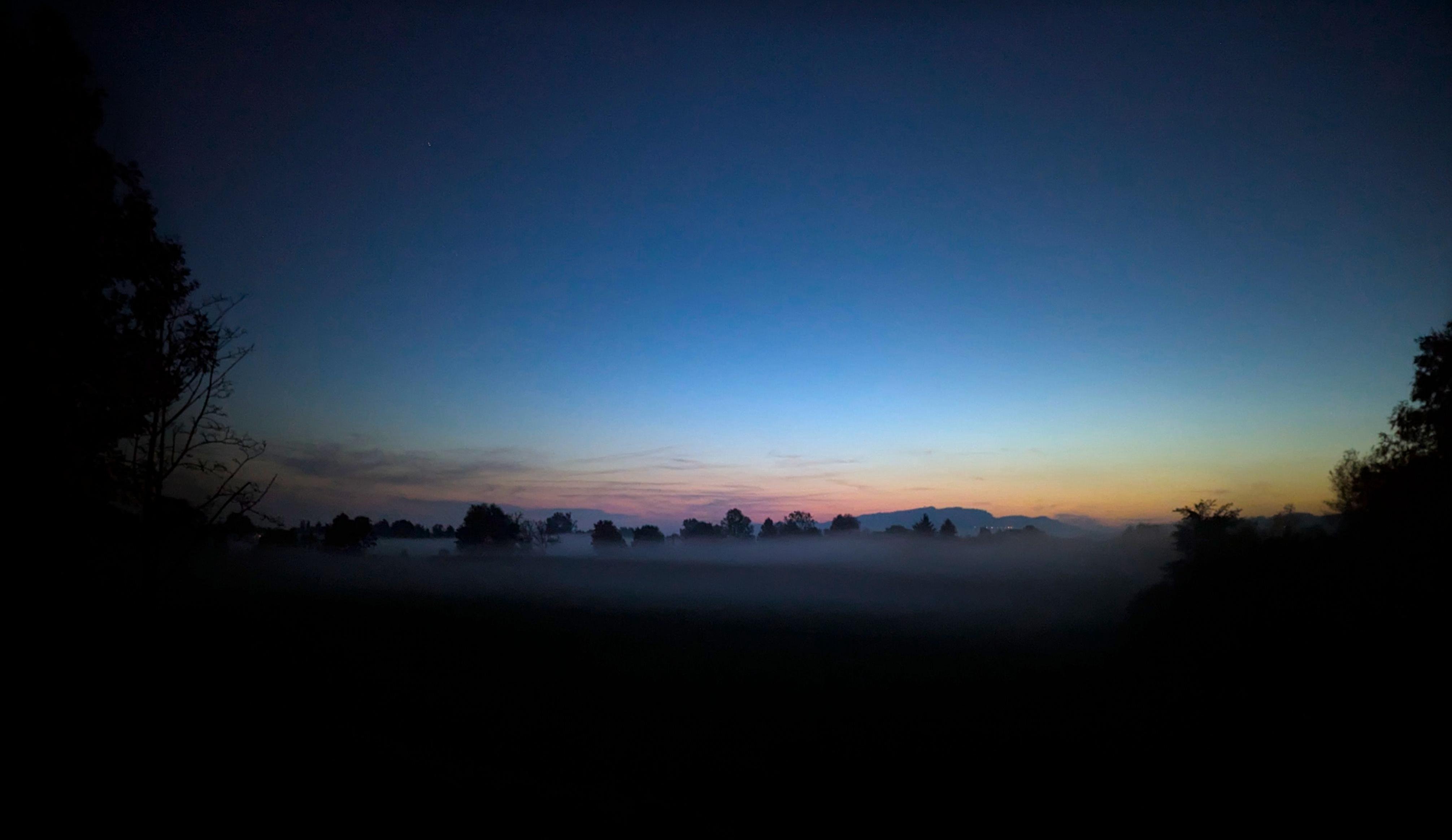 Wide landscape at dawn with a blue-to-orange gradient sky, a low band of fog across the horizon, and dark silhouettes of trees and distant hills framing the scene.