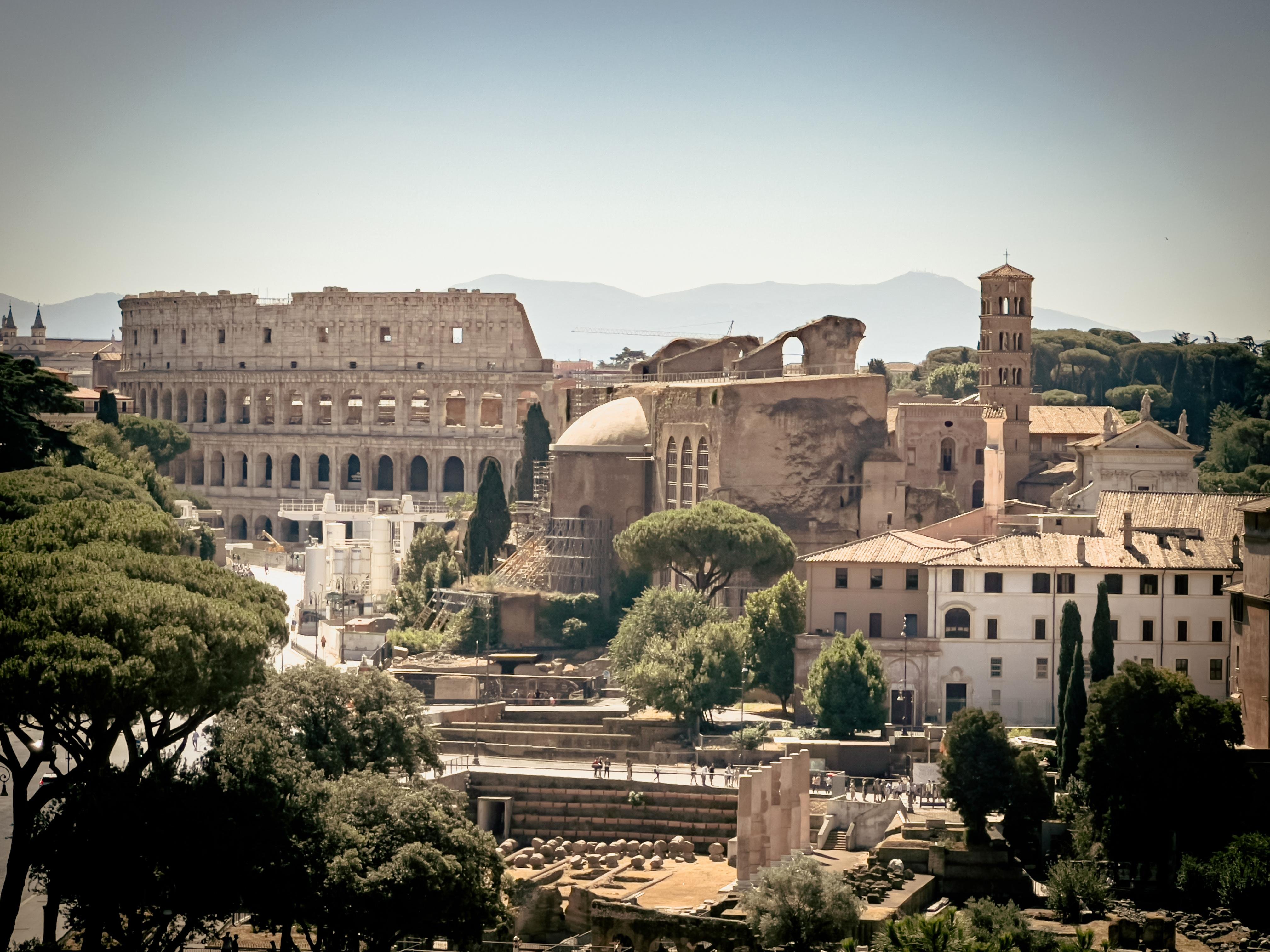 Colosseo a Roma, Italia. È un antico anfiteatro romano e uno dei monumenti più iconici al mondo. Intorno al Colosseo ci sono vari edifici e strutture storiche, con alberi e vegetazione. Sullo sfondo, si vedono colline o montagne distanti.