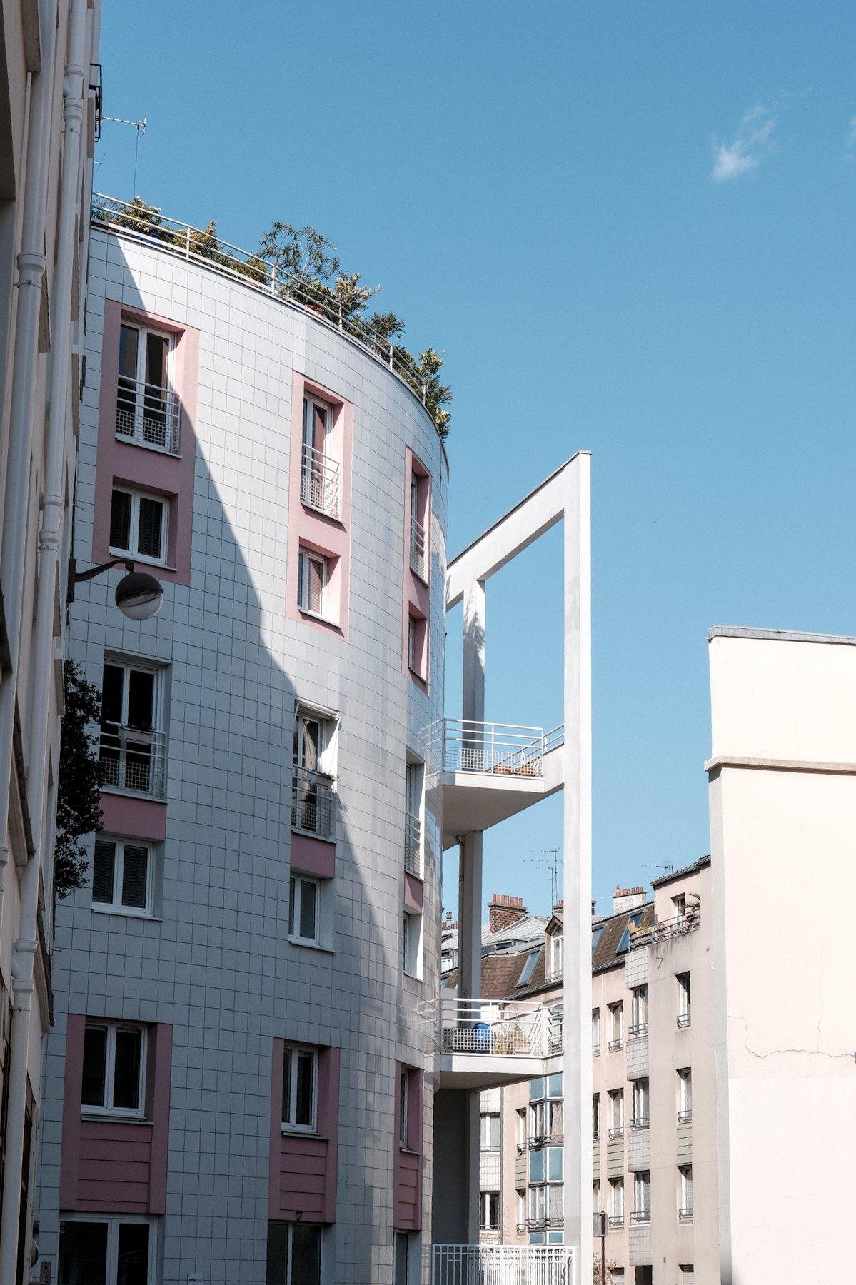 Photo verticale. Sur la gauche, un immeuble cylindrique couvert de carrelage blanc, avec les bords des fenêtres en rose pâle, sous un ciel bleu avec juste un tout petit nuage en haut à droite