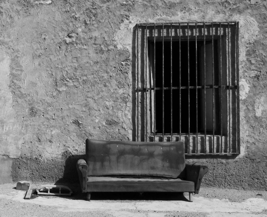 Fotografía en blanco y negro en la que aparece una pared exterior de una casa en ruinas. En la pared, en el lateral derecho de la foto, hay una ventana grande y abierta con rejas. Bajo la ventana, levemente a la izquierda y en el centro de la foto, hay un sofá viejo, sucio y harapiento pegado a la pared. A la izquierda del sofá hay unos ladrillos.