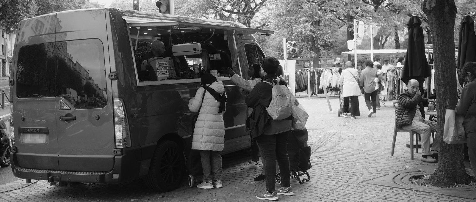 Photo paysage noir et blanc montrant la camionnette d’un serrurier, avec des clients devant. On voit aussi un bout de terrasse de café avec un homme assis à une table, et des gens au fond qui marchent entre les étals d’un marcher