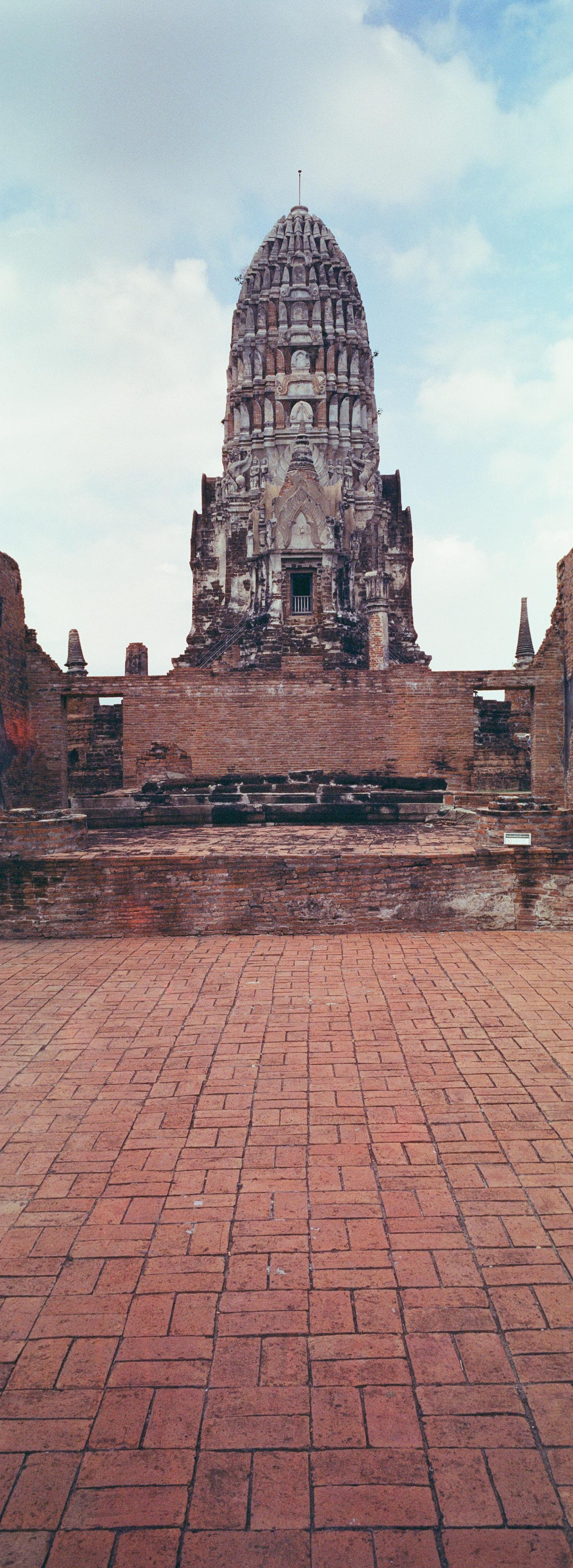 Panoramic color photo, portrait orientation, of a tall stone buddhist temple tower centered behind low red-brick ruins and paving, in front of a cloudy blue sky filling the background