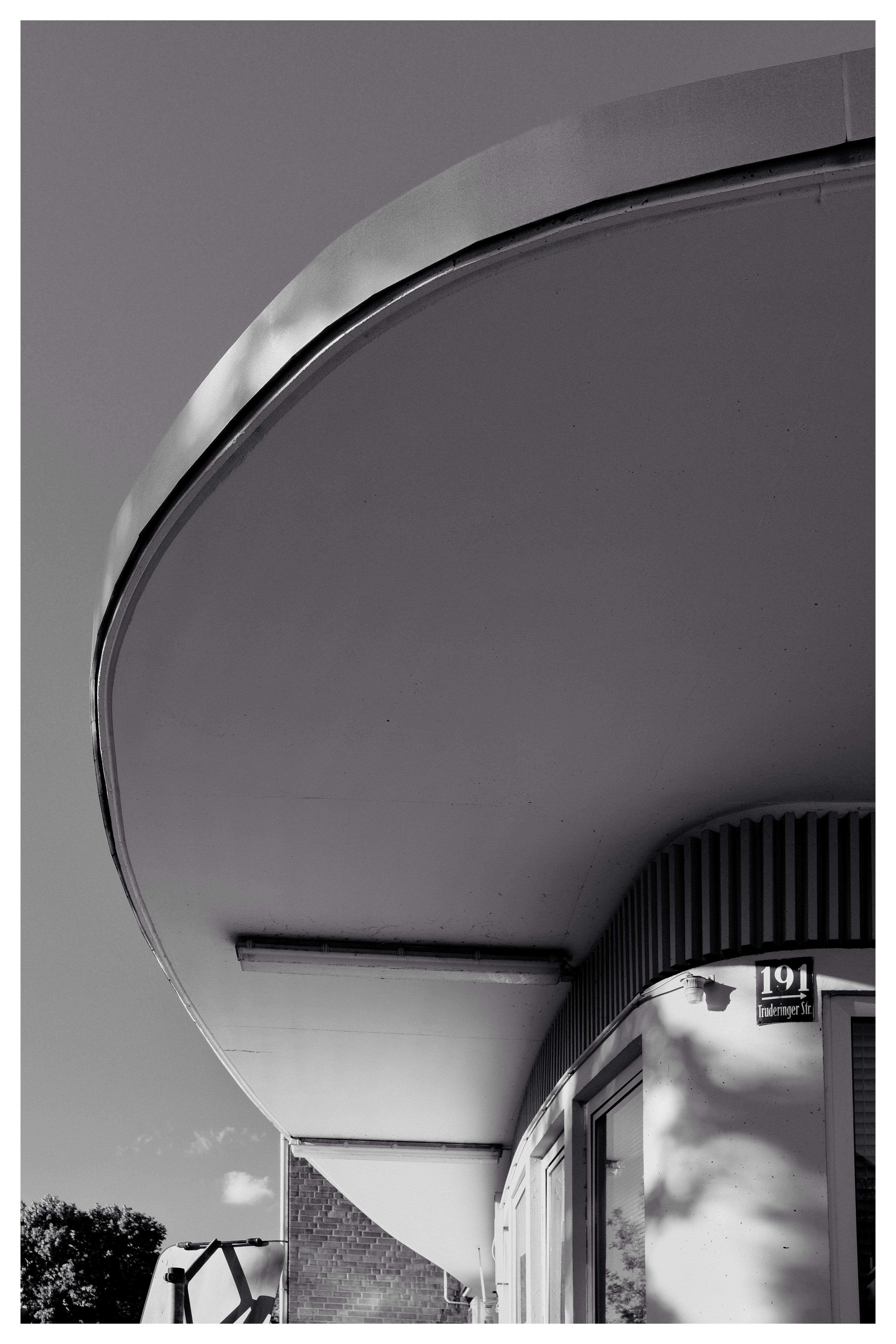 Curved, overhanging roof structure dominates the upper half of the image. Below, a brick building facade features windows and vertical stripes. Bottom left shows a partial view of a tree and vehicle in the background. Black and white color scheme.