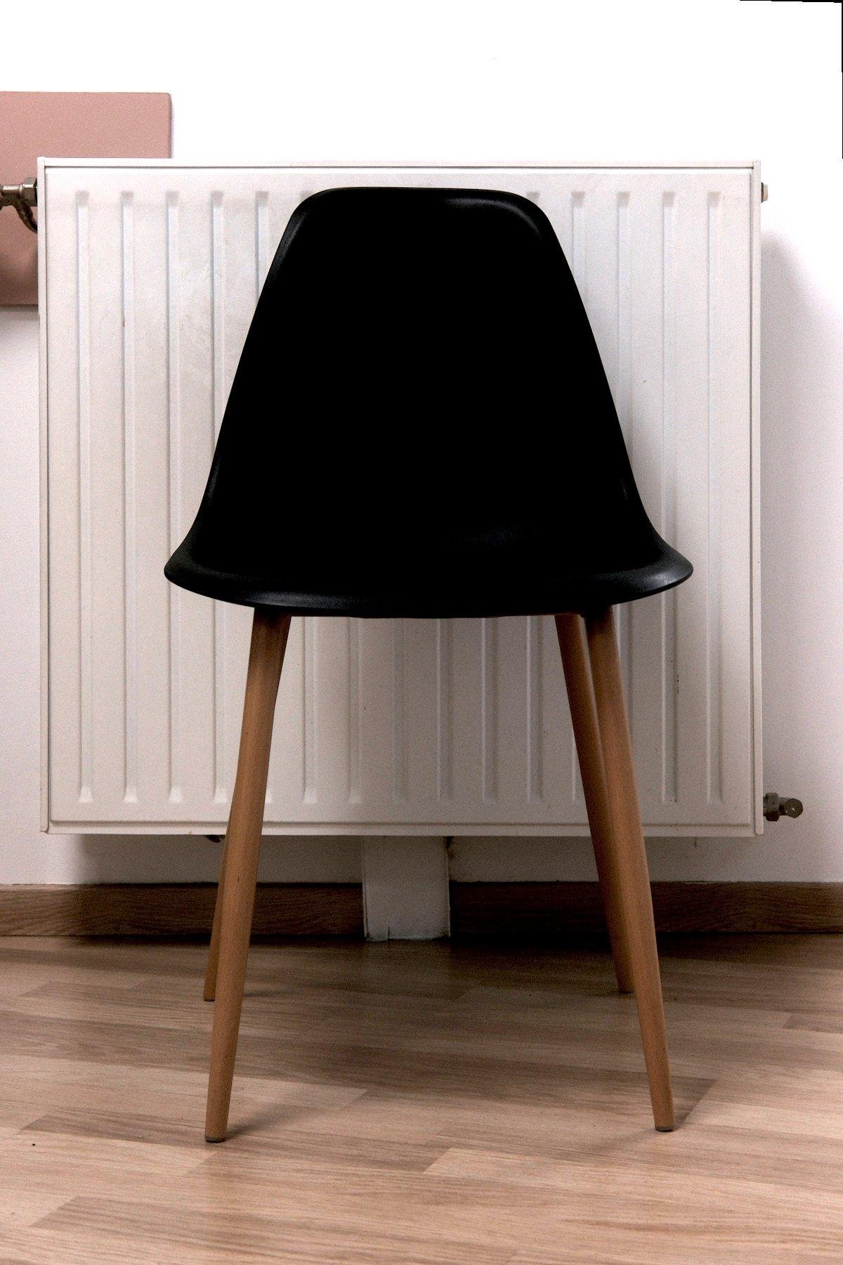 Photo verticale. Une chaise design mais en plastique bas de gamme noir sur des pieds en bois assez fins posés sur un plancher, devant un radiateur blanc et carré