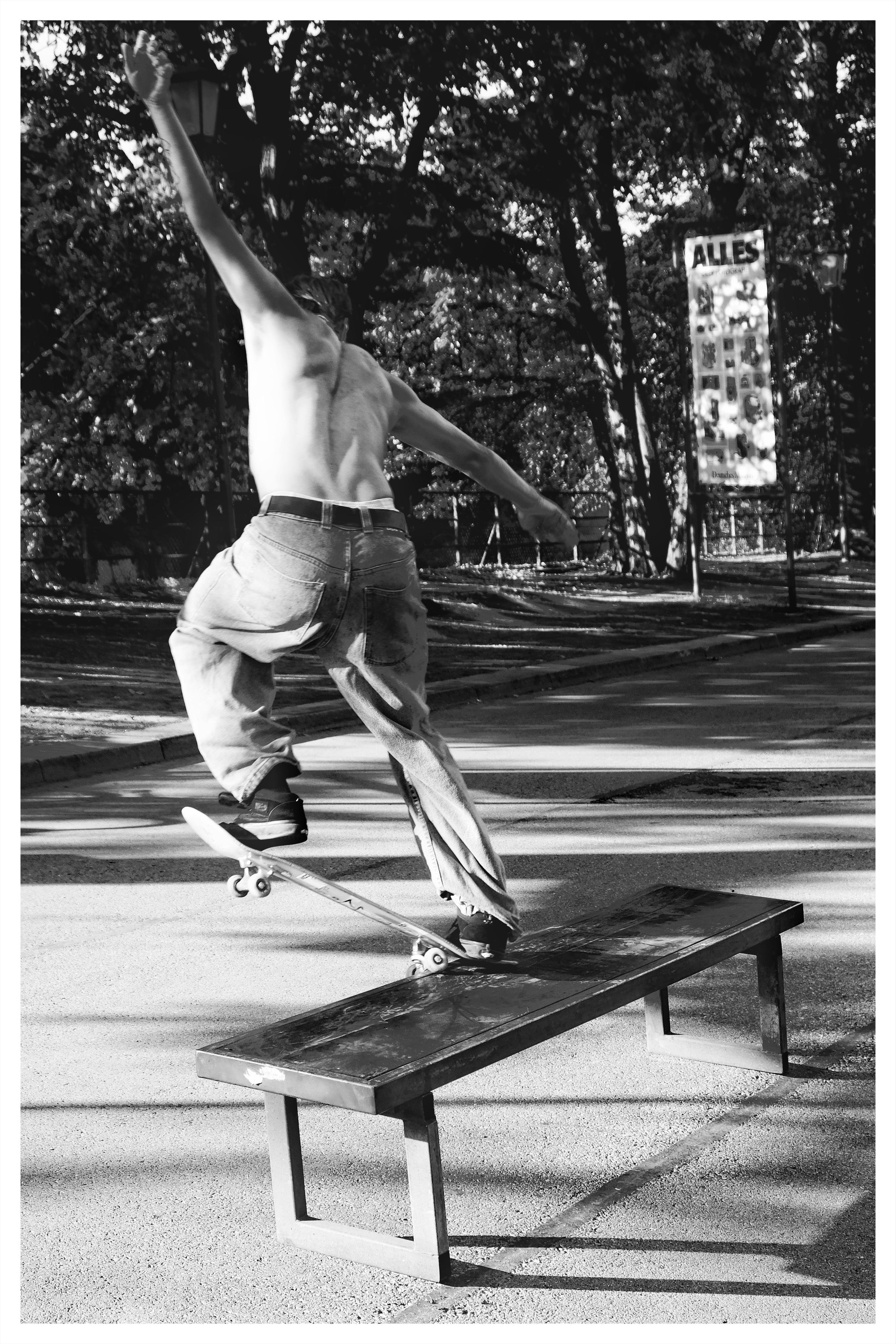 Black-and-white photo of a shirtless skateboarder in baggy jeans balancing on a skateboard as it slides along a low metal grind box in a park, with trees and a street sign in the background.