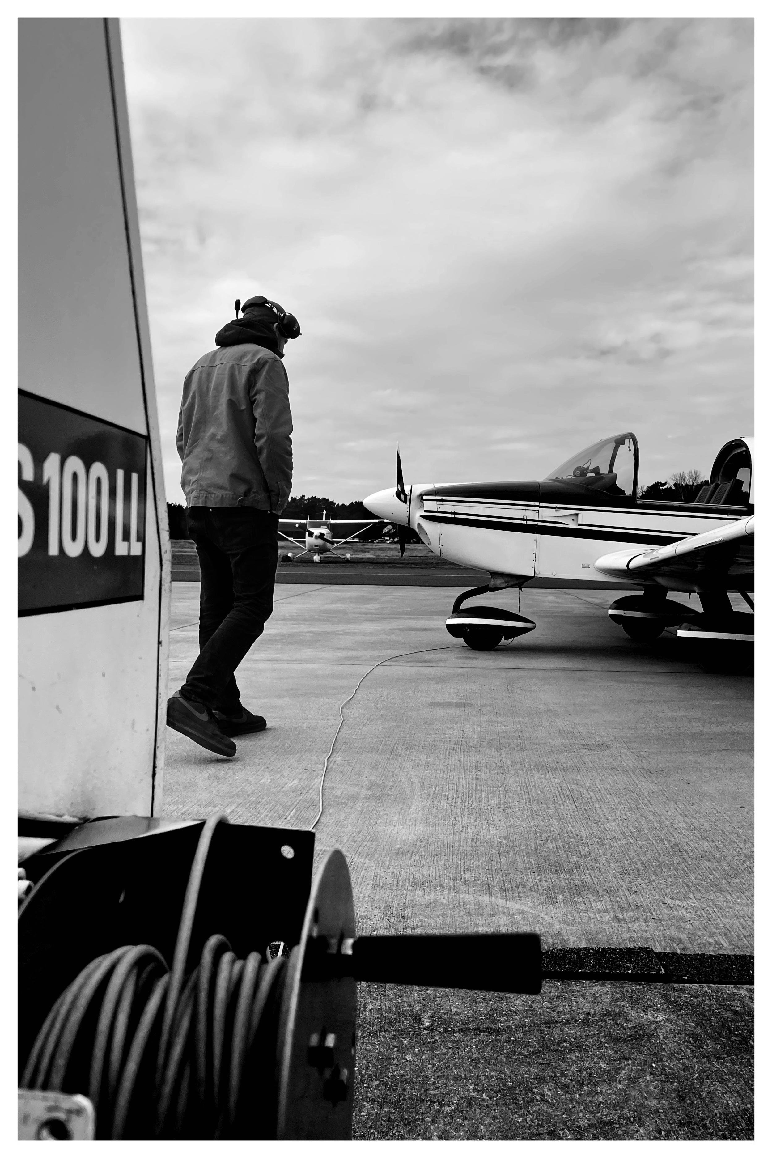 Person wearing a headset walks across an airport tarmac toward a small single‑engine propeller airplane with its canopy open; a cable reel and the edge of another aircraft or equipment frame the left foreground under a cloudy sky.