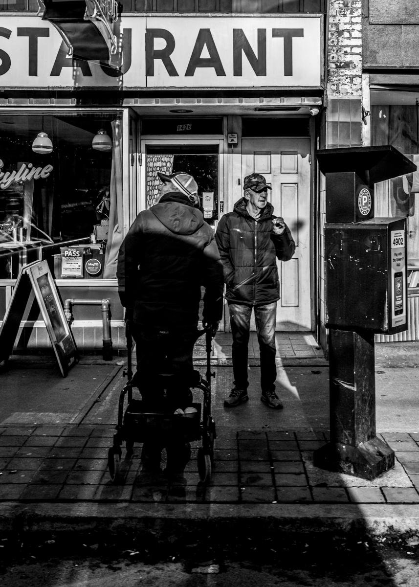 Black-and-white street scene outside a storefront with a large “RESTAURANT” sign; two men in jackets stand on the sidewalk, one pushing a small cart or stroller, the other facing the camera holding an item, with a parking meter at the right and a window display and sandwich-board sign at the left.