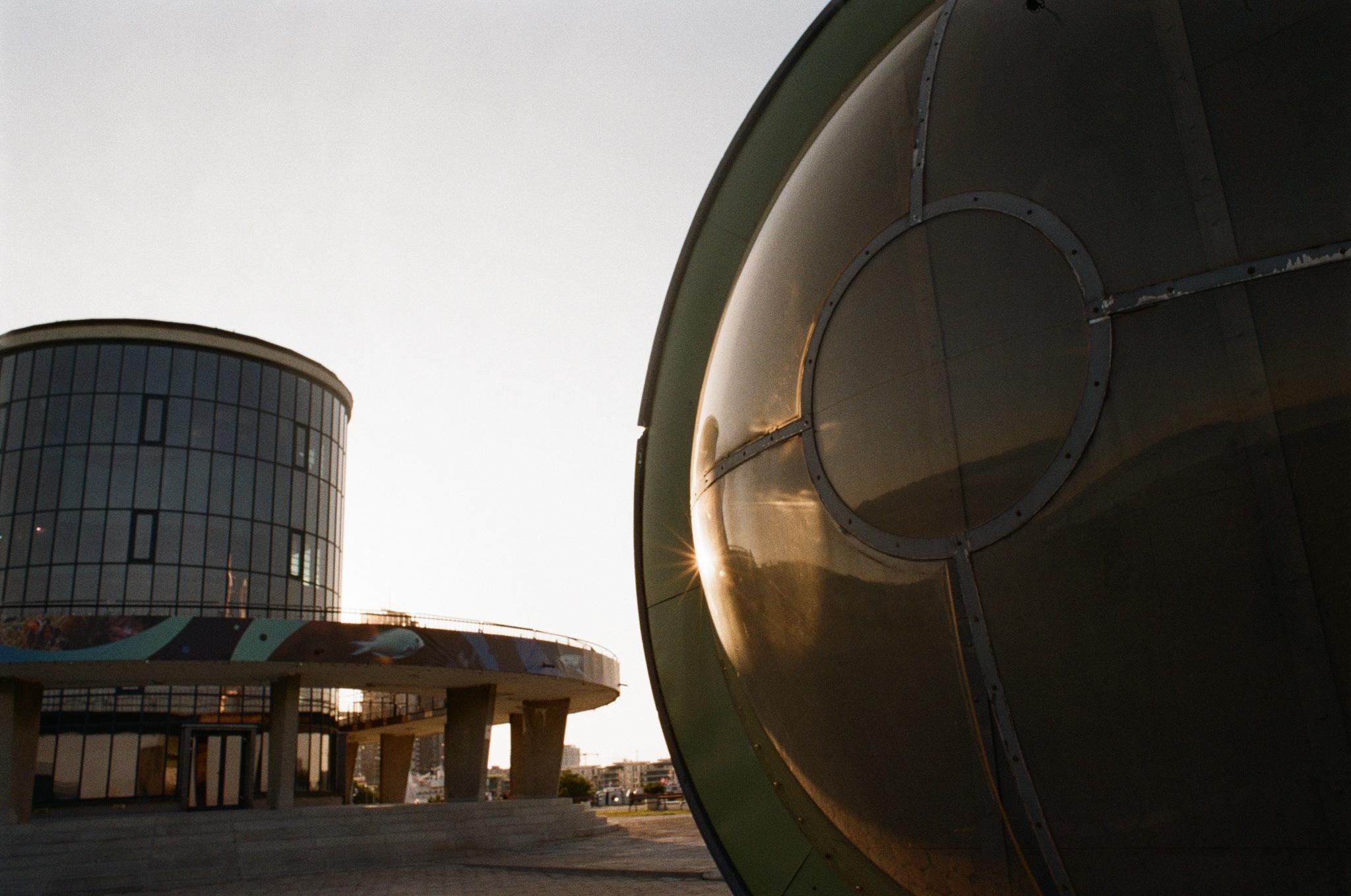 Large metallic spherical structure in the right foreground, partially cropped, with sunlight reflecting off its surface; modern cylindrical glass building with a circular raised platform and columns on the left under a pale sky.