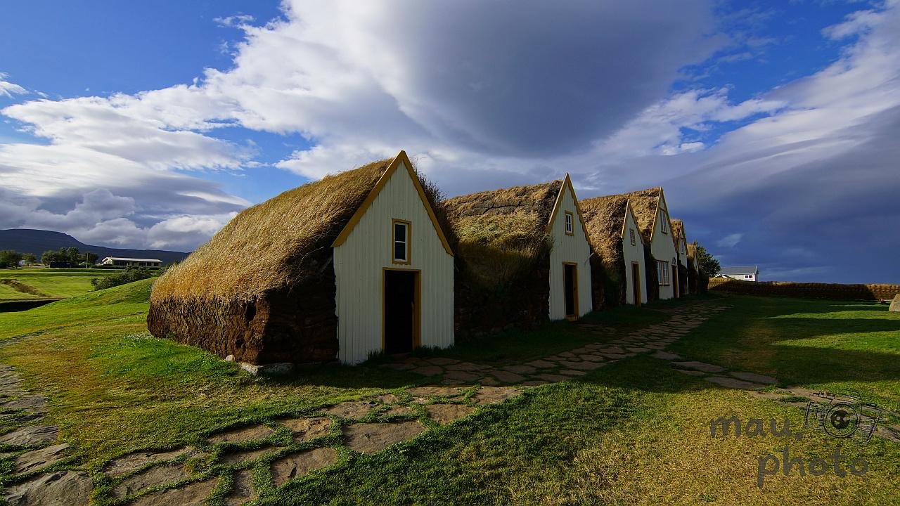 Row of small white gabled houses with thick turf roofs along a stone path in a grassy field, under a blue sky with large layered clouds.