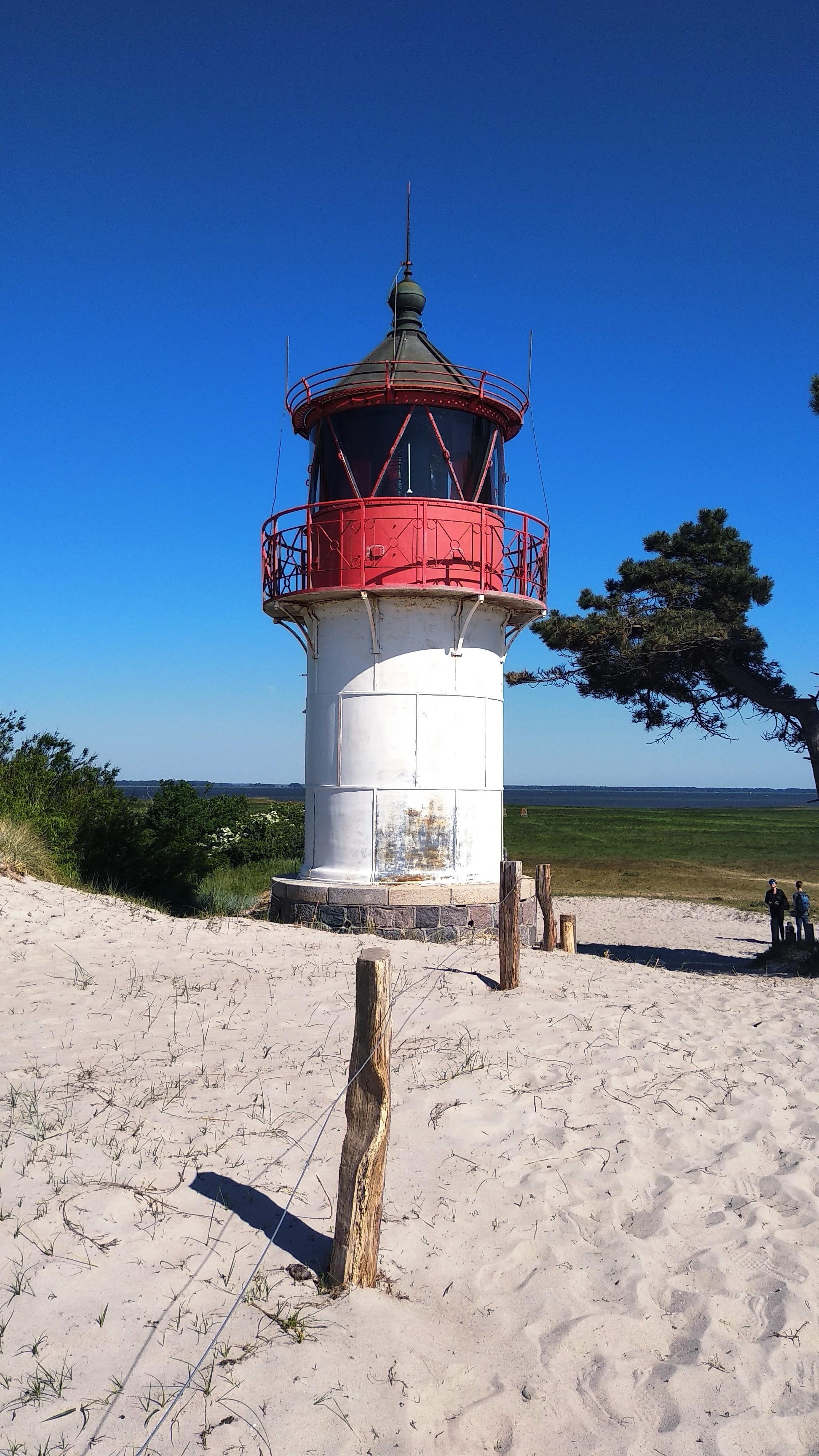 Red and white lighthouse on a sandy hill, surrounded by sparse vegetation. Clear blue sky in the background. A wooden fence is visible in the foreground, along with a single tree to the right. Two people are standing in the distance near the water.