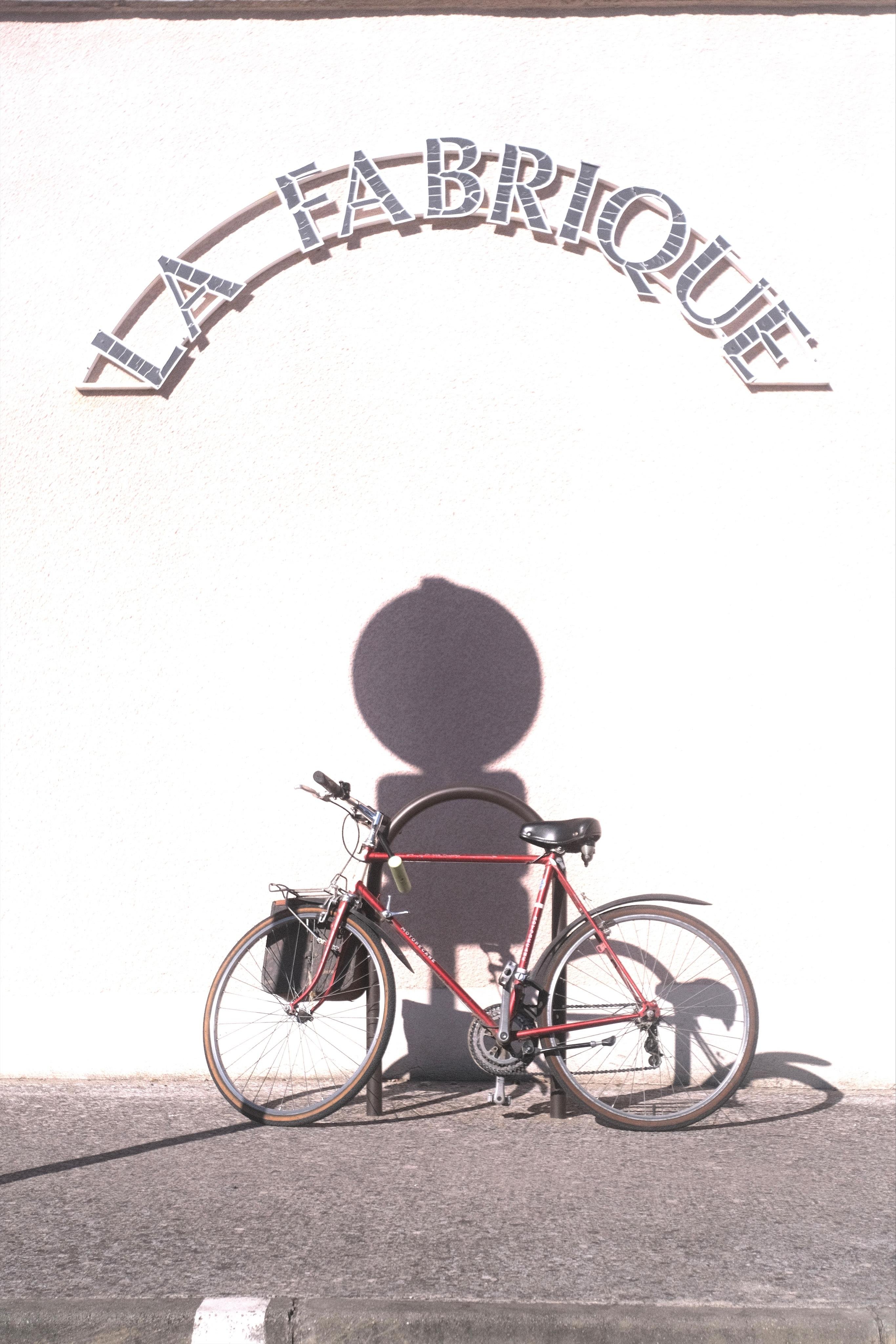 Red bicycle locked to a bike rack in front of a wall overexposed in sunlight, with the arched wall sign “La Fabrique” above and a street sign shadow cast on the wall.