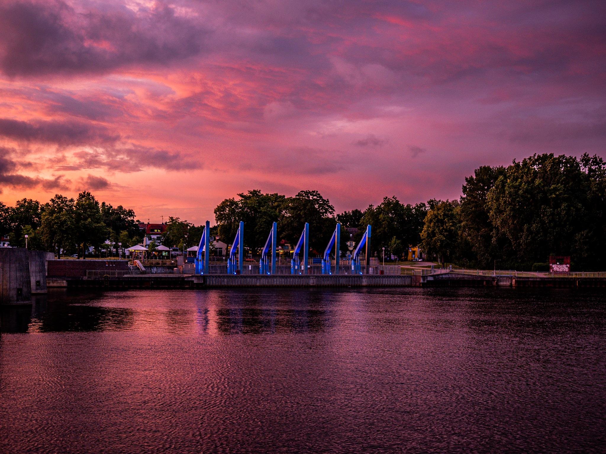 A waterfront scene at sunset with a vibrant pink and purple sky. Blue architectural structures resembling sails are positioned along the edge of a waterway. Trees and buildings are visible in the background, partially silhouetted against the colorful sky. The water reflects the hues of the sunset, adding a warm glow to the scene.