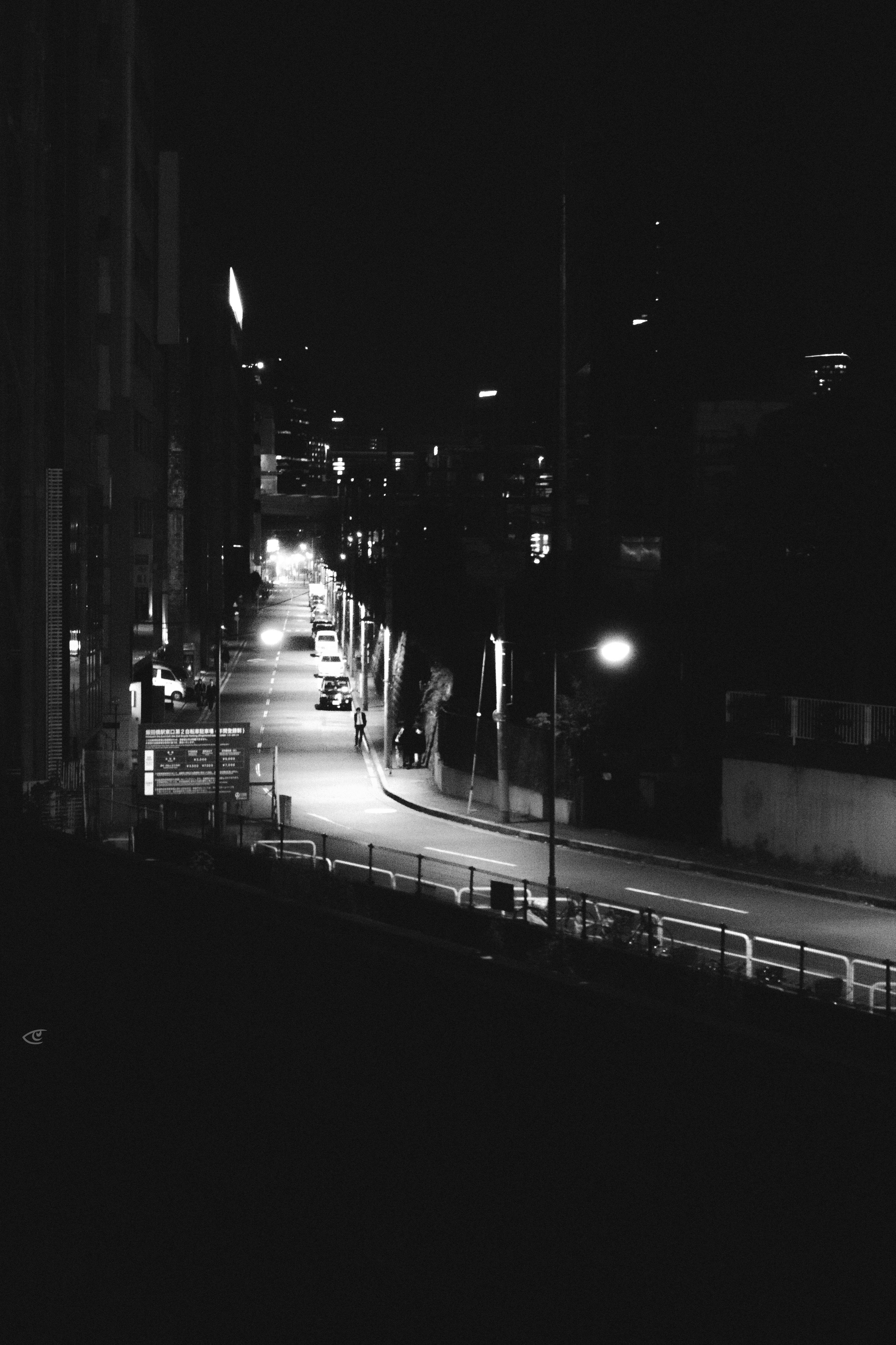 Black-and-white nighttime view down a long city street, with streetlights illuminating the road, parked cars along the curb, a few pedestrians on the sidewalk, and tall dark buildings framing the scene.