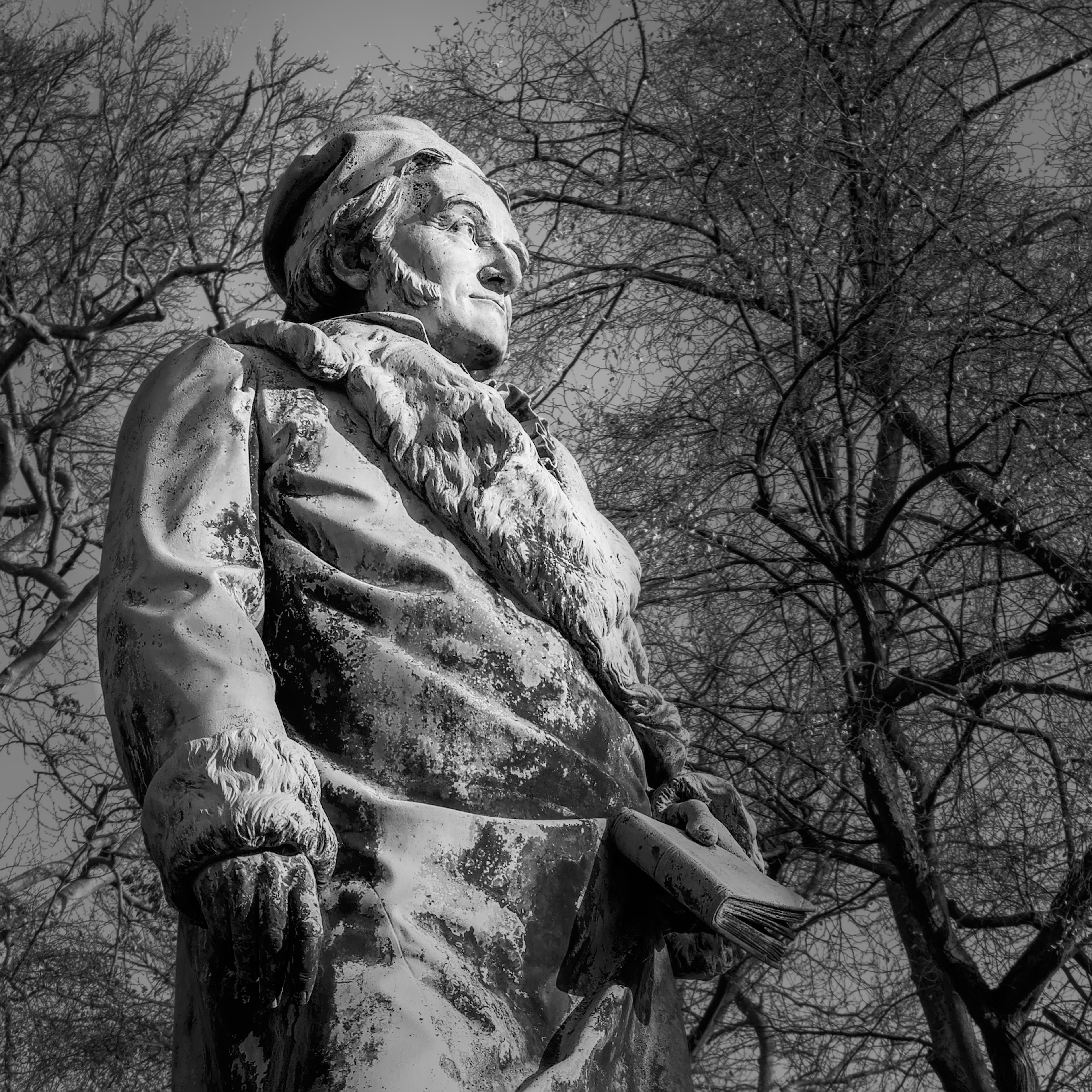 Low-angle black-and-white photo of a weathered stone statue of a man in a fur-trimmed coat and cap, holding a  book at their side, with bare tree branches filling the background sky. 
### 
Schwarz-weißes Low-Winkel-Foto einer verwitterten Steinstatue eines Mannes in einem mit Fell beschnittenen Mantel und Mütze, der ein Buch an seiner Seite hält, mit nackten Baumzweigen, die den Hintergrundhimmel füllen. 