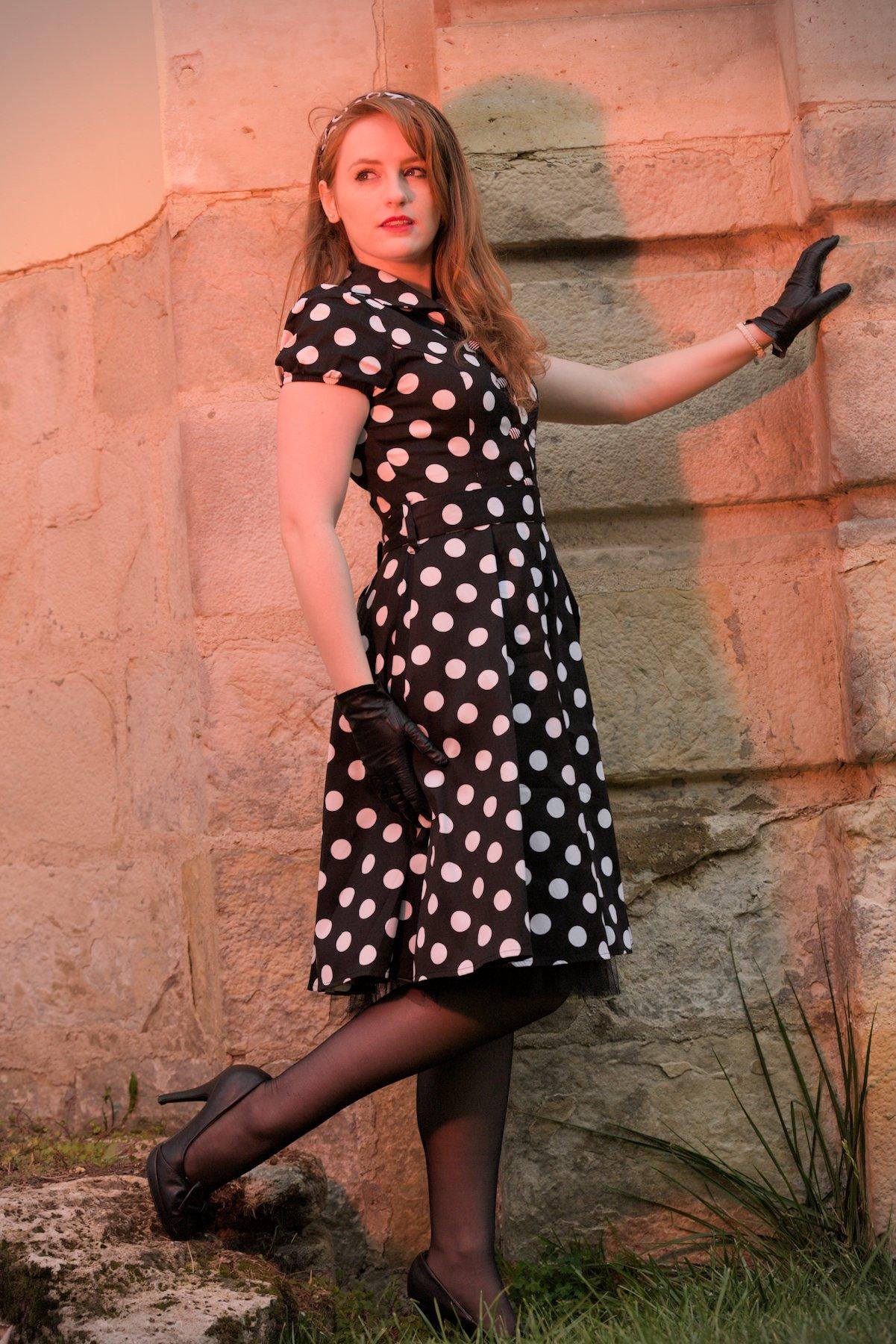 Photo verticale. Une jeune femme aux cheveux long roux, posant devant un mur de vieilles pierres, portant une robe noire à pois blanc, des collants noirs, des chaussures noires avec un petit nœud, des gants  en cuir noir et un serre tête noir avec des motifs blancs. Elle descend la dernière marche d’un petit escalier en vielles pierres, la main gauche se tenant à un mur, en regardant sur sa droite