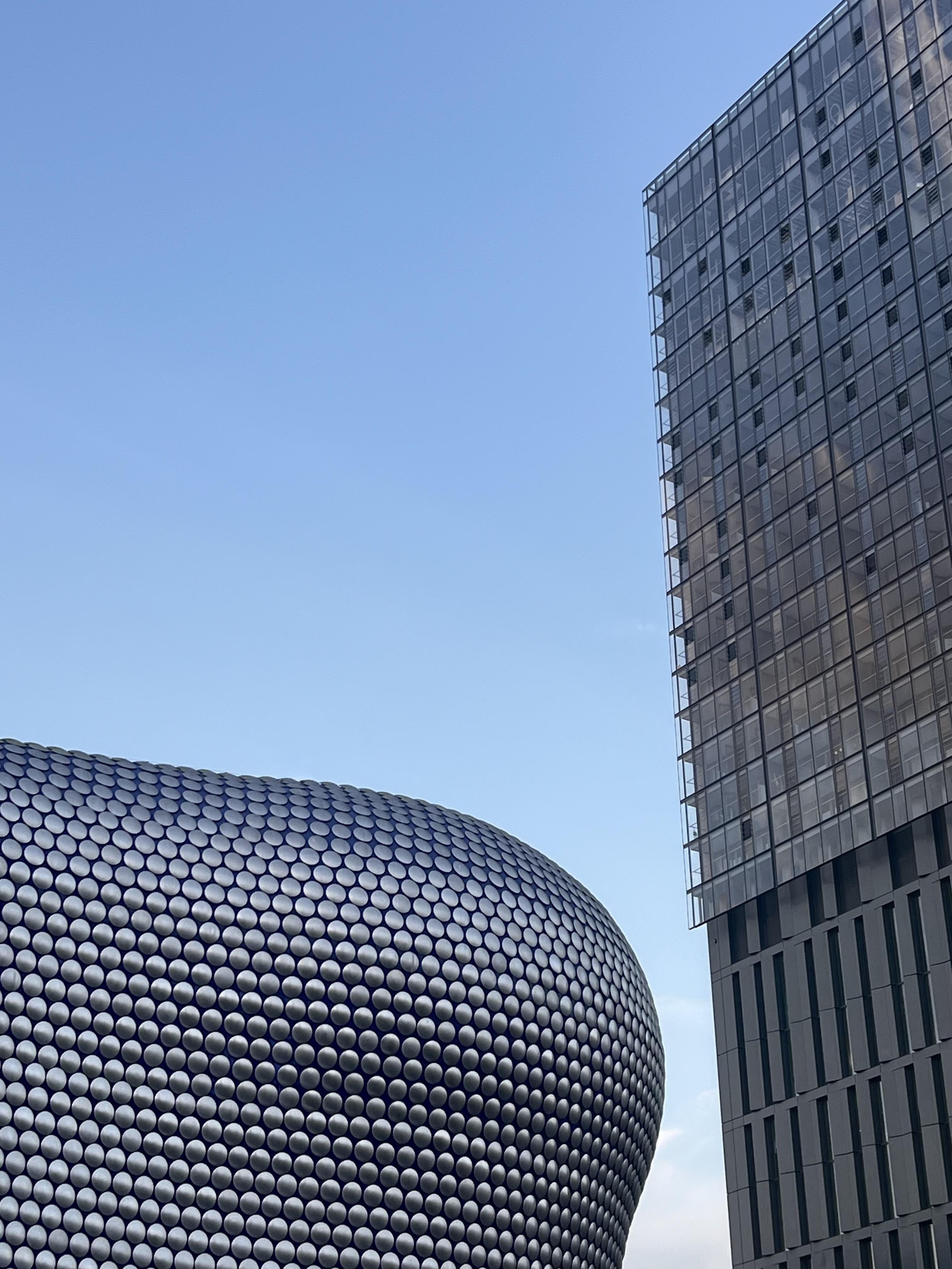 The curved Selfridges building in Birmingham, which is covered in circular metallic discs is on the left of photo, with the tall, angular glass-and-steel Beorma Tower on the right against a clear blue sky.