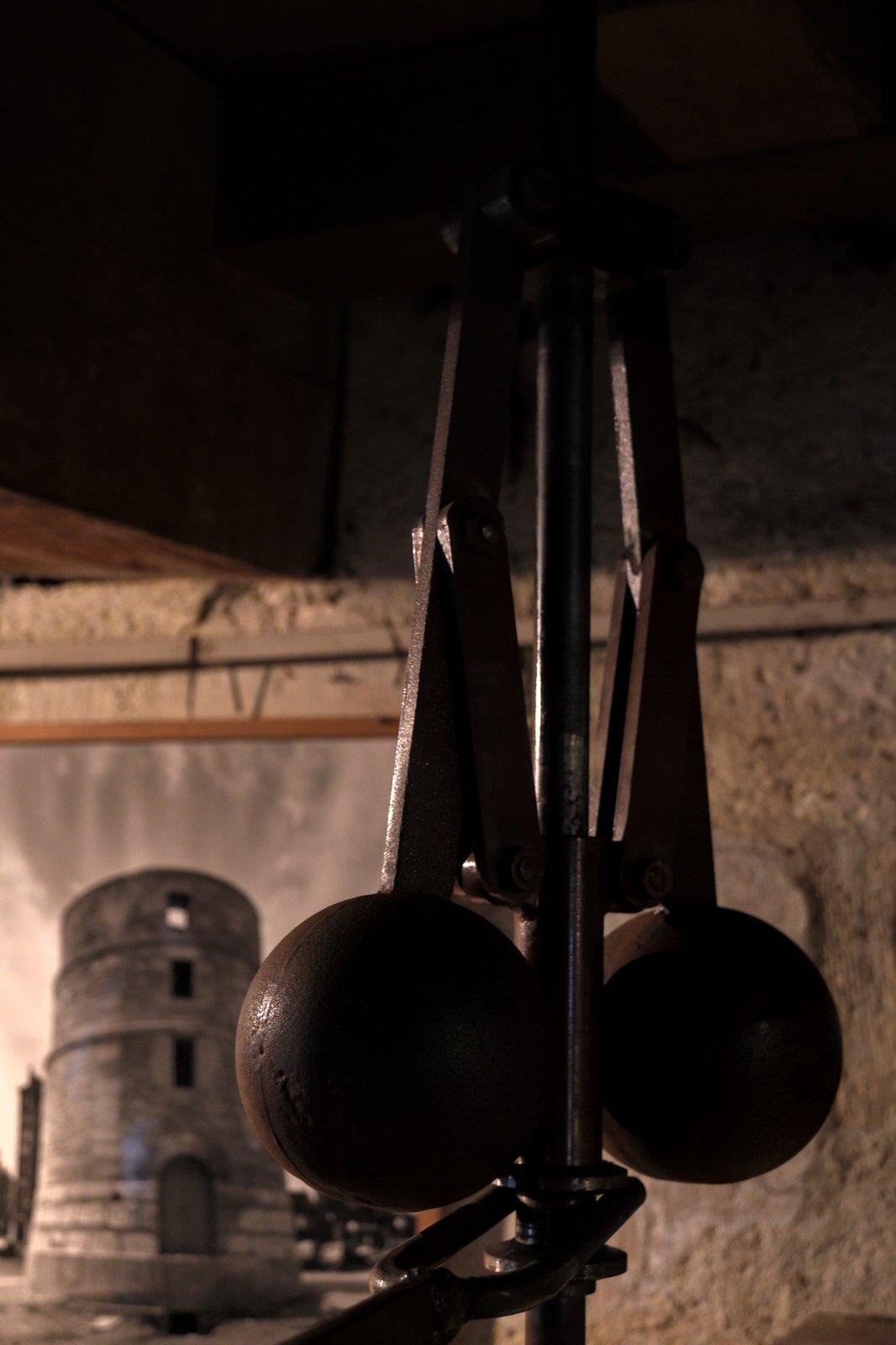 Photo verticale. Morceau d’un mécanisme constitué de de 2 bras en métal accrochés à une tige centrale, terminés par des poids sous forme de sphères. On devine que cet ensemble fait partie d’un tout hors cadre.