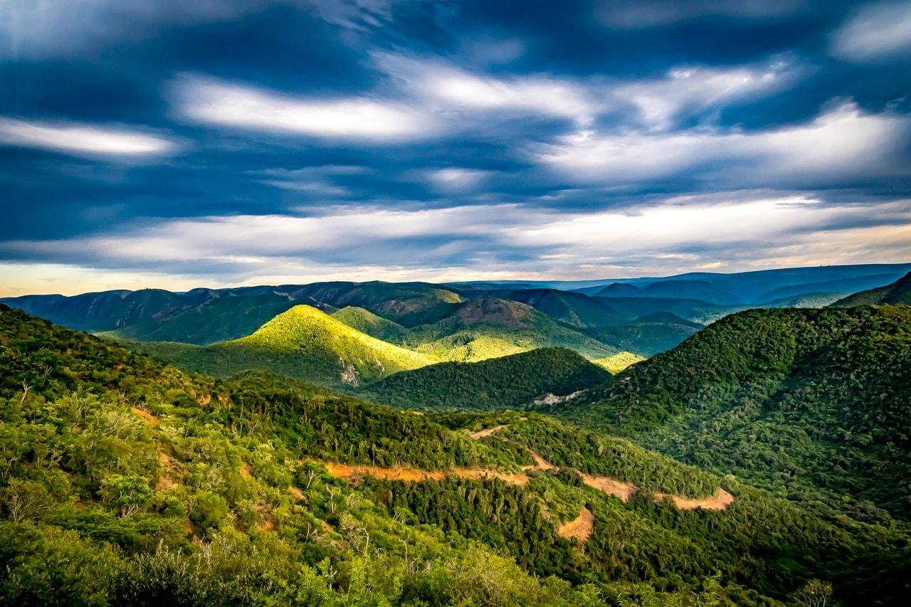 The image depicts a scenic view of a landscape with lush green mountains and valleys under a dramatic sky. The clouds are moving, creating patches of sunlight across the hills. The overall atmosphere is serene and picturesque.