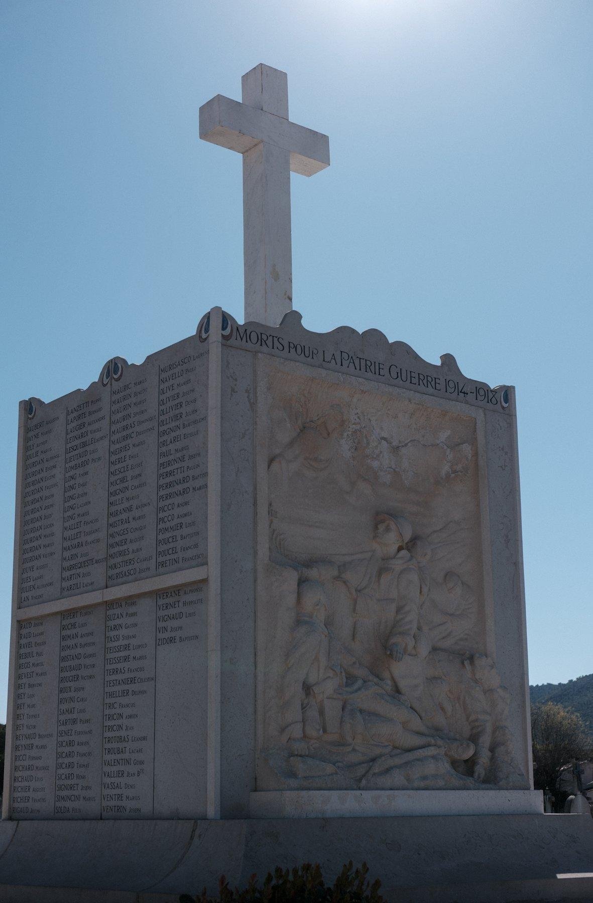 Photo verticale. Un monument à la mémoire des soldats tombés lors de 14-18. Un grand cube avec sur une face, des noms, une autre un relief sur le thème de la guerre, surmonté d’une croix blanche.