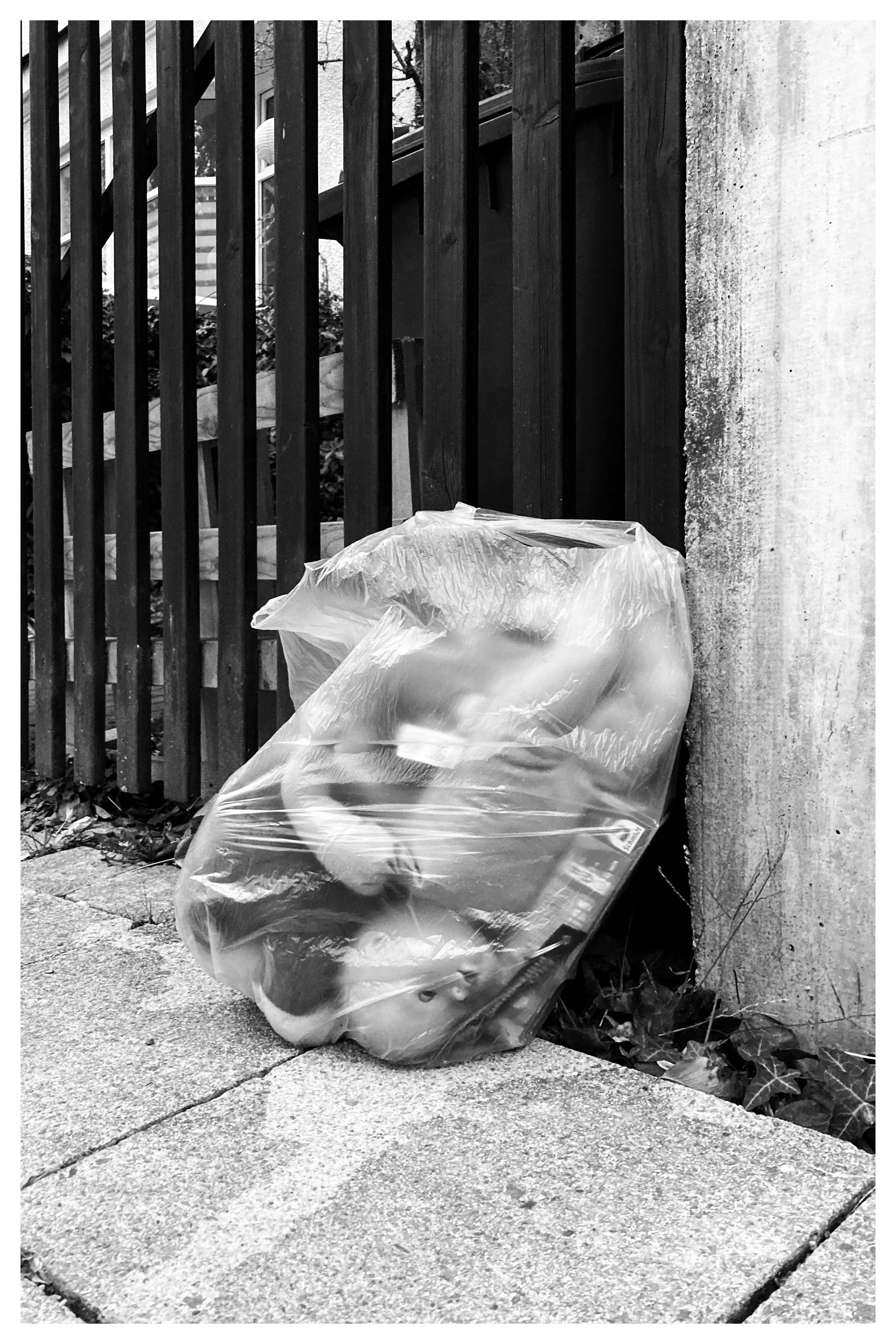 Clear plastic trash bag filled with soft items, including a visible plush toy face, leaning against a concrete post beside a vertical wooden fence on a paved sidewalk.