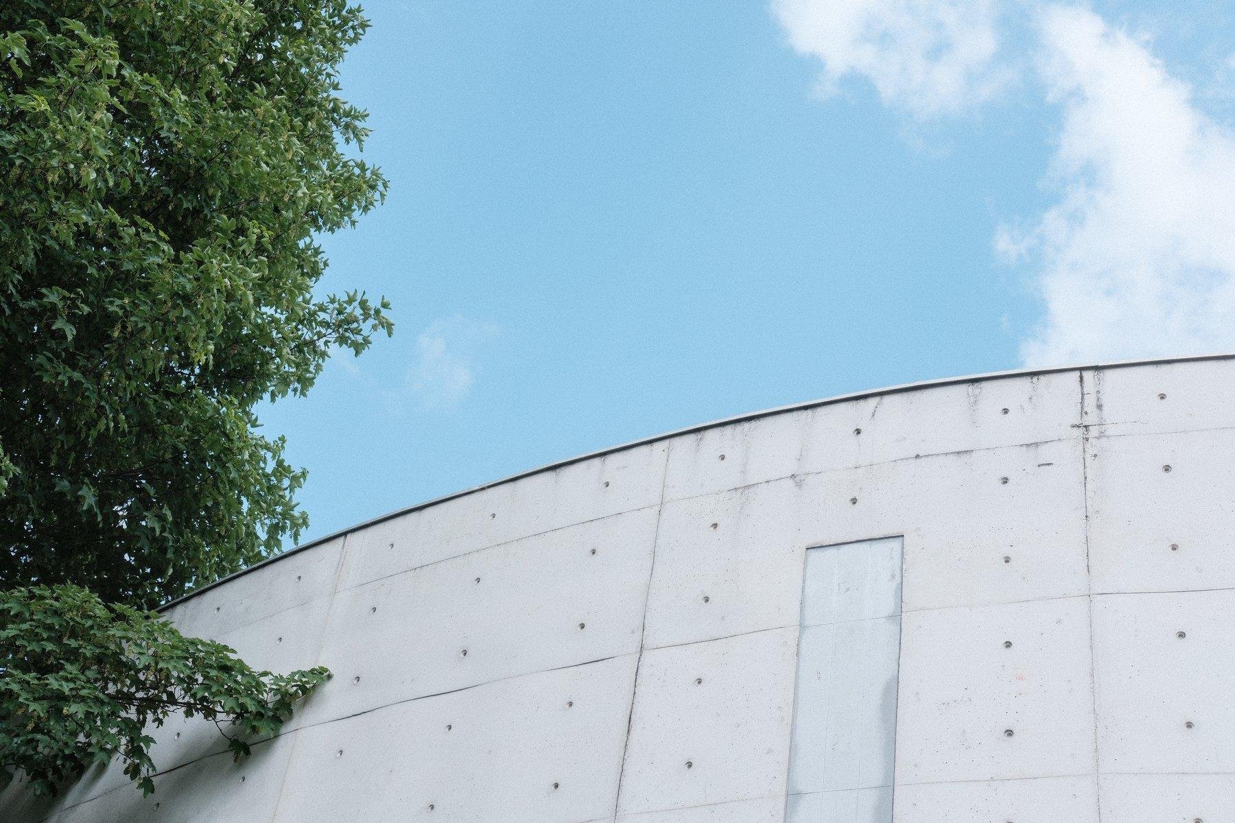 Photo horizontale. Contre plongée sur un morceau de bâtiment circulaire d’un étage de hauteur, blanc avec des trous régulier sur les dalles constituant le mur. À gauche, le feuillage de printemps d’un arbre et au dessus, le ciel bleu avec un nuage blanc